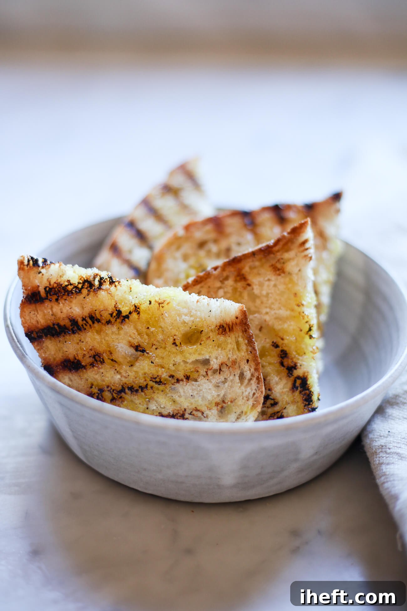 Classic Fettunta Tuscan Bruschetta in a grey ceramic bowl on a marble counter. The Tuscan slices of bread have grill marks and are coated in a drizzle of olive oil.