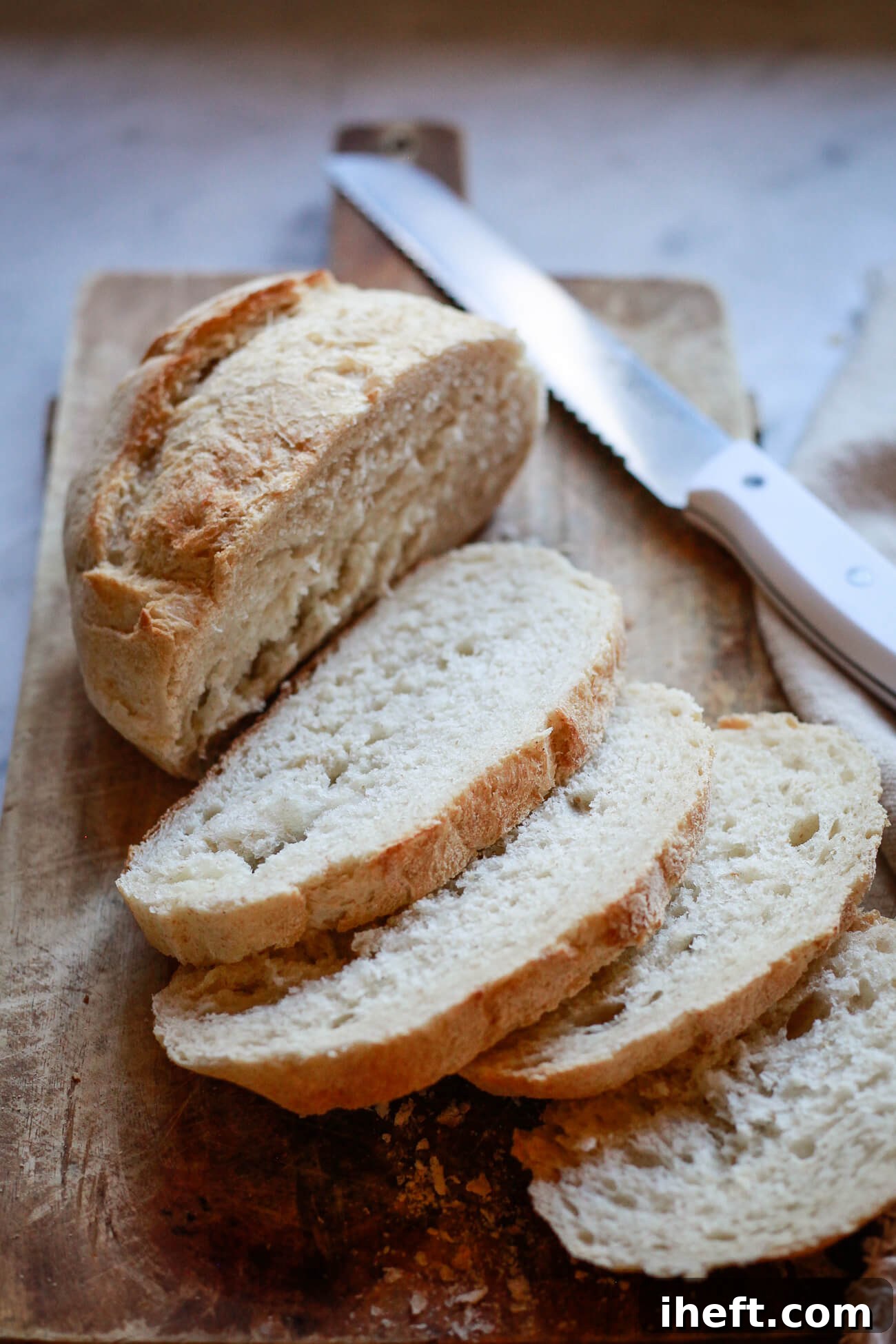 A rustic loaf of Pane Toscano or Tuscan Bread, half sliced, on a wooden cutting board with a serrated knife.