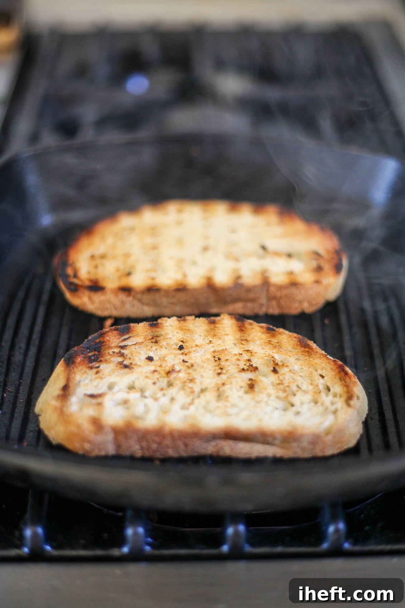 Two pieces of Tuscan bread with grill marks continue to cook on a black grill pan to make fettunta bruschetta.