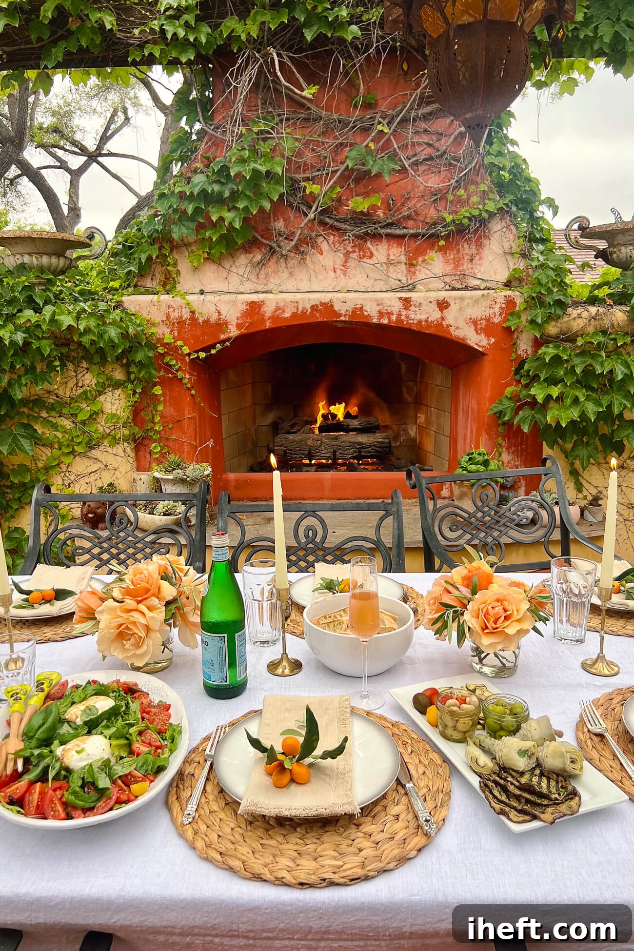 A beautiful al fresco Italian dinner party in front of an outdoor fireplace. The table is set with a white linen tablecloth, and family-style plates of food are on the table including a Burrata Salad and antipasti platter. A beautiful Italian dinner party idea. 