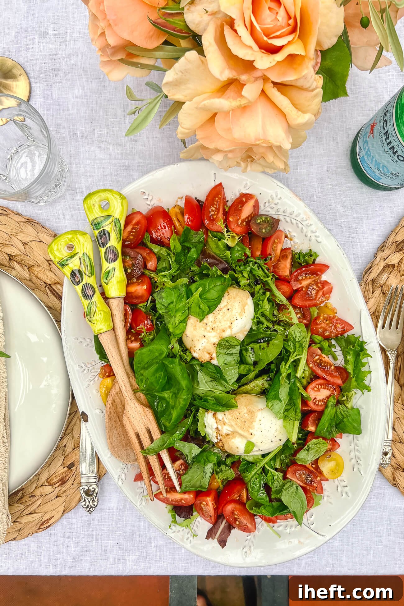 A beautiful large white ceramic platter with mixed greens topped with tomatoes and two balls of burrata cheese. This is a great Italian dinner party menu salad served on white tablecloth with peach colored roses. 