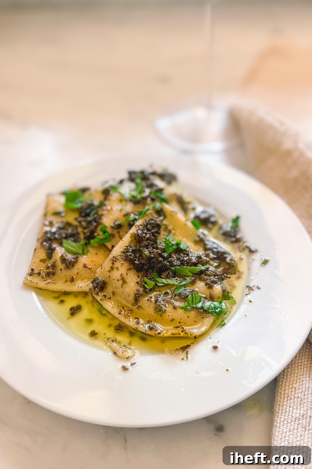A white plate with ravioli covered in black truffle sauce garnished with parsley. 