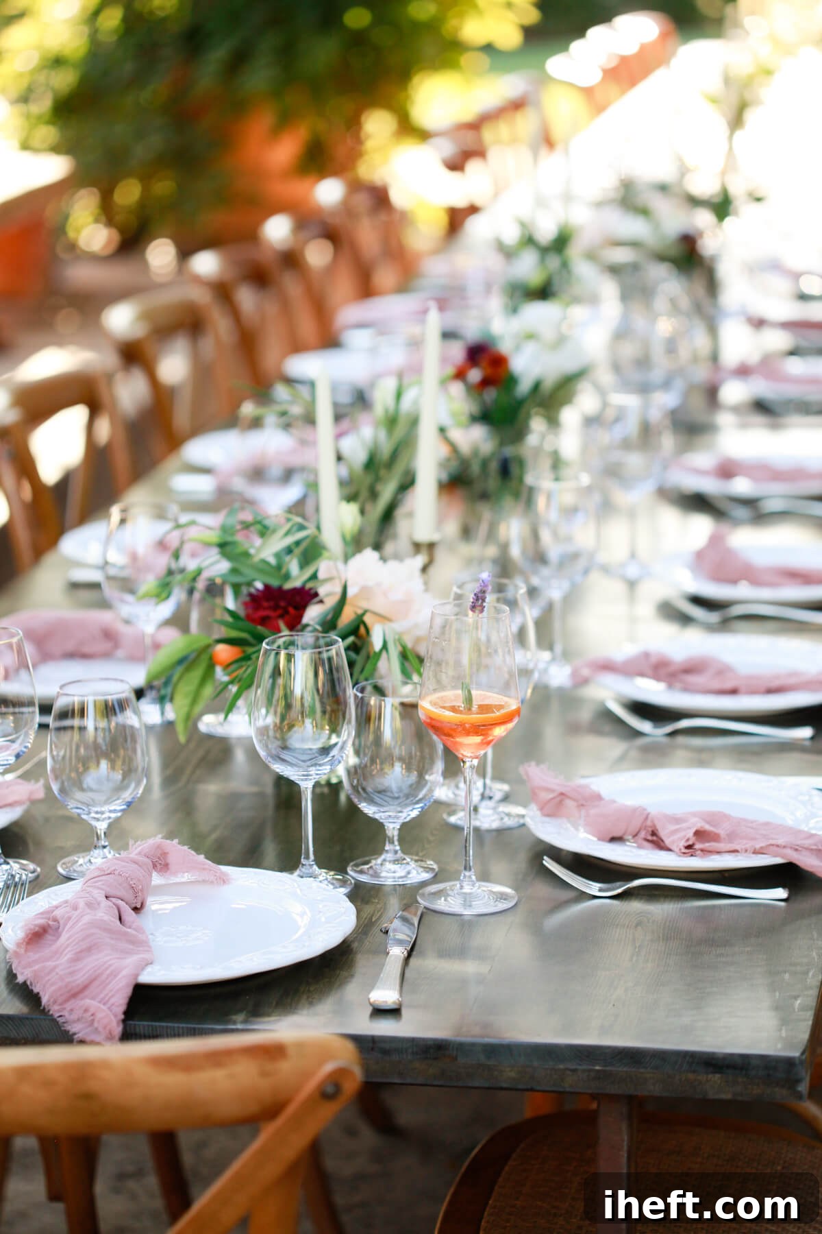A beautiful long table set in a backyard. An aperol spritz sits on the table set with white dishes, pink napkins, and olive branch floral arrangements. 