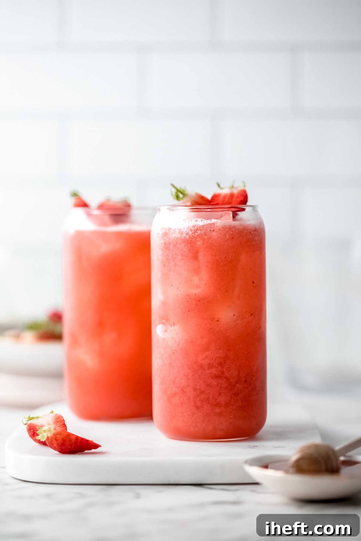 Two tall glasses of vibrant red strawberry agua fresca, garnished with fresh strawberry slices, sitting on a clean white kitchen counter.