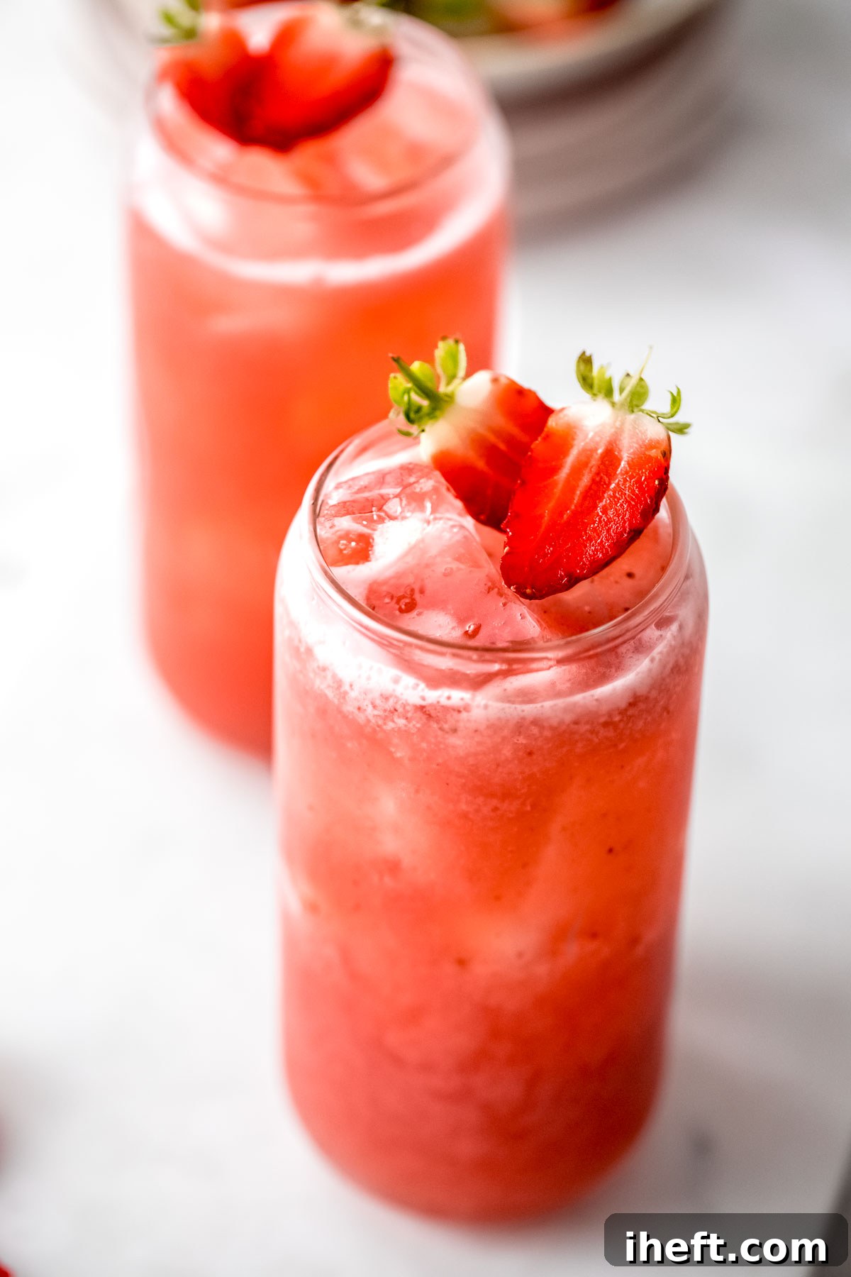 Two clear glasses filled with ruby-red agua de fresa (strawberry water), brimming with ice cubes and adorned with fresh strawberry halves, set against a blurred background.