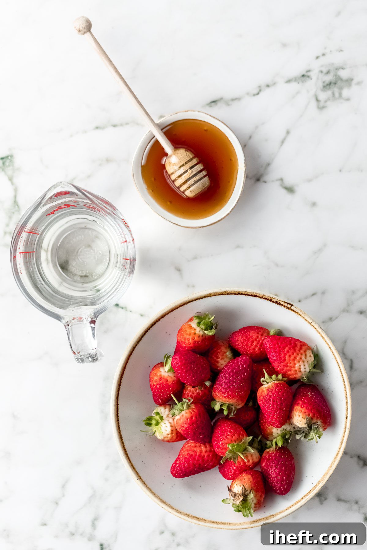 A minimalist flat lay photograph displaying the core ingredients for agua de fresa: a small ceramic bowl of golden honey, a vibrant bowl of ripe red strawberries, and a glass pitcher of clear water, all arranged on a smooth marble countertop.