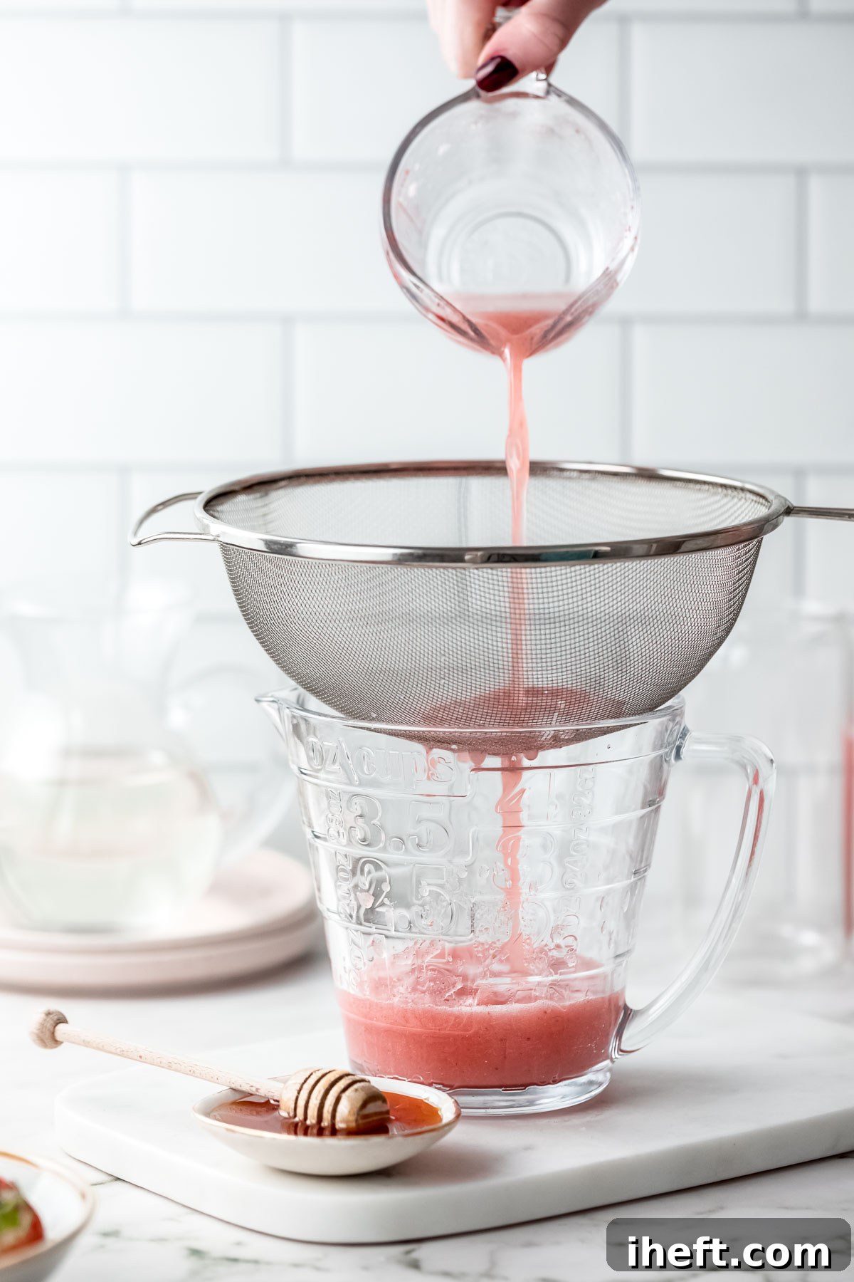 A close-up shot of vibrant red strawberry water being carefully poured through a fine-mesh sieve into a large glass bowl, which rests on a pristine marble counter. This step illustrates the straining process of making strawberry agua fresca.