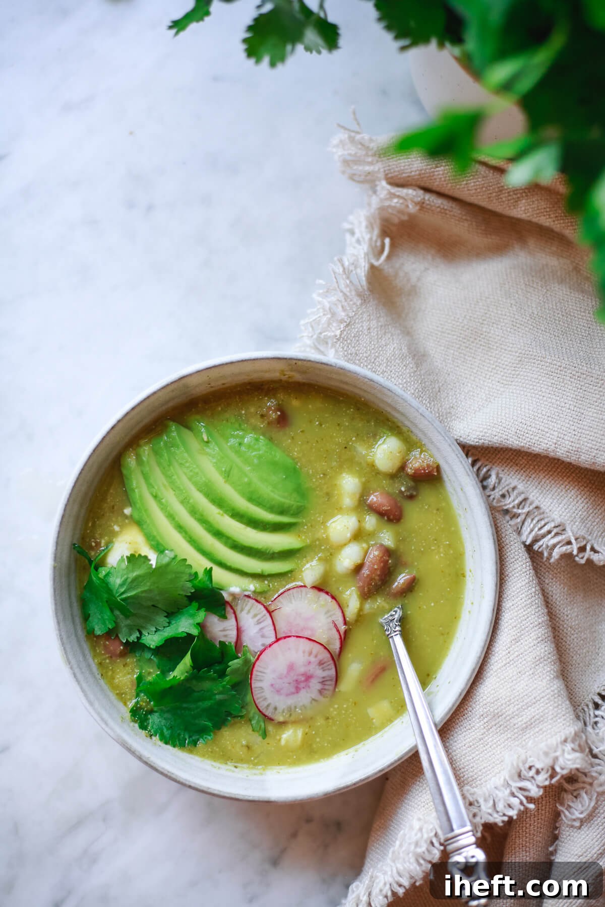A bowl of vegetarian pozole verde with pinto beans in a grey bowl with silver spoon. The pozole is garnished with avocado, radish, and cilantro.