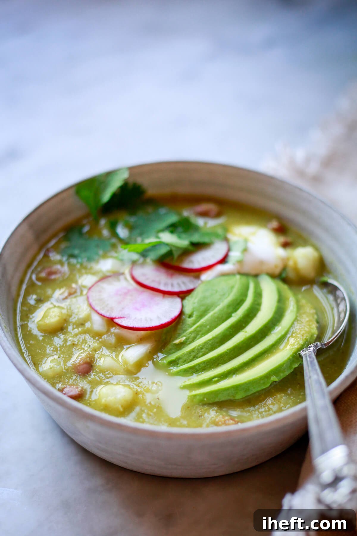 A bowl of vegan pozole in a grey ceramic bowl. A Mexican pinto bean and hominy soup topped with avocado, radish slices, and cilantro.