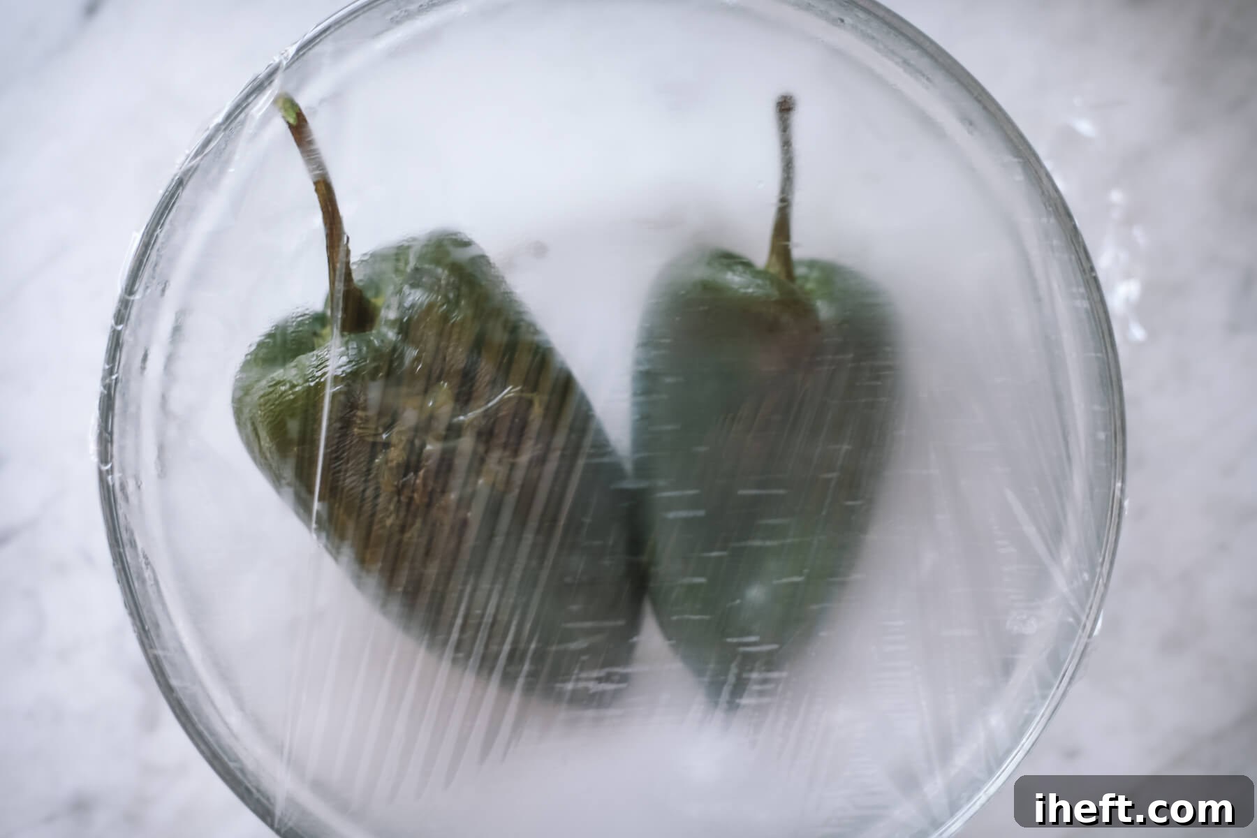Two poblano peppers steam in glass bowl covered with plastic wrap.