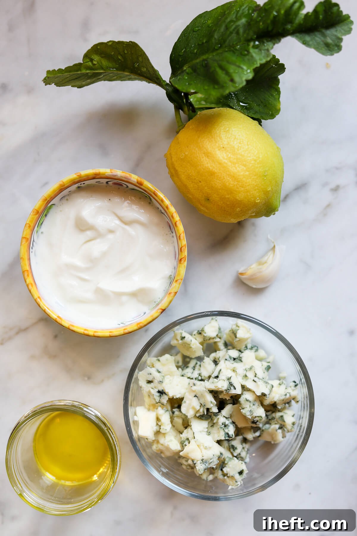 The ingredients for Blue Cheese dressing sit on a marble counter. Lemon, garlic clove, crumbled Roquefort cheese, plain yogurt in a bowl, and a little olive oil in a small dish. 