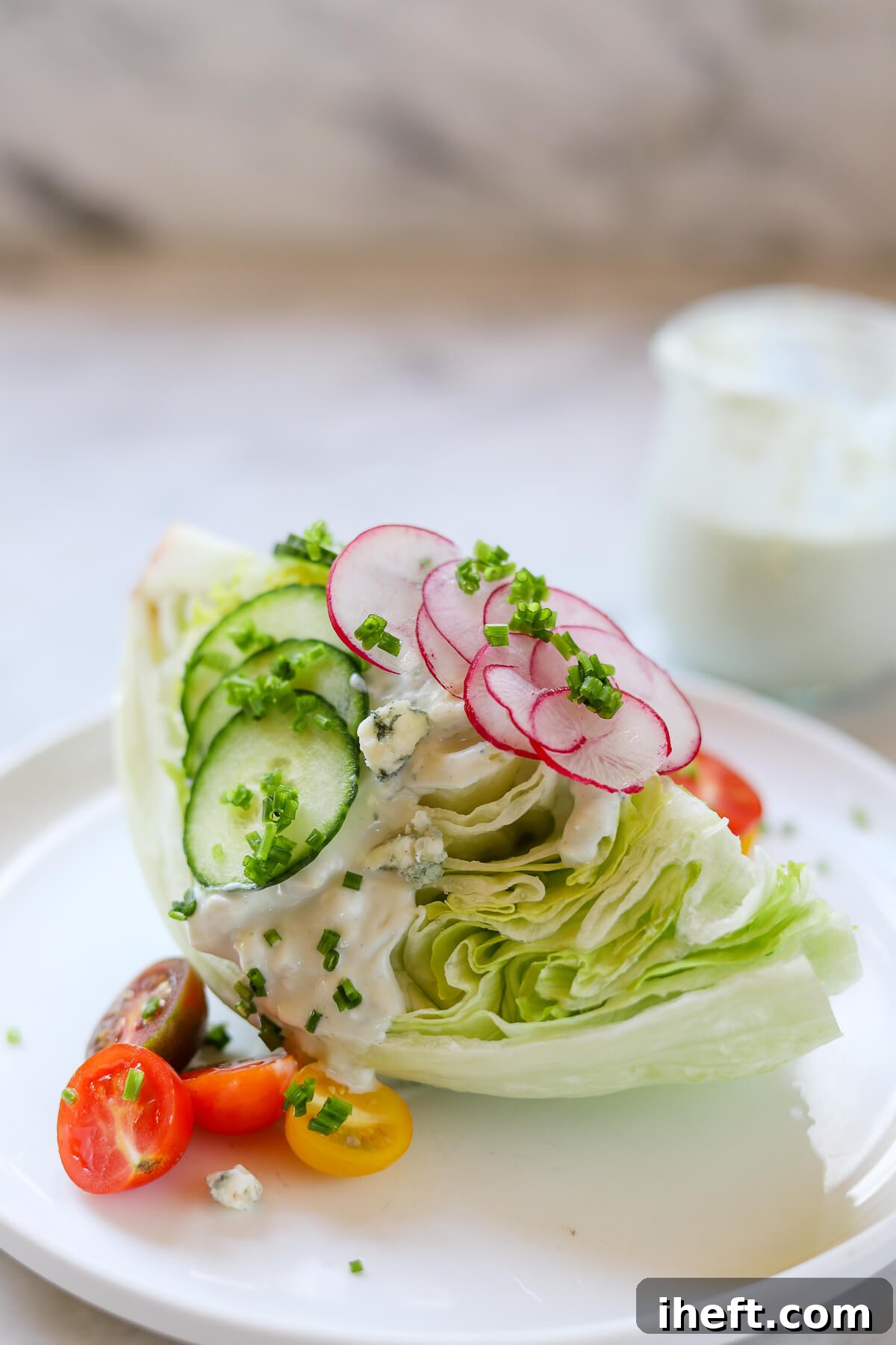 A wedge salad topped with Roquefort Dressing, cucumber and radish slices, halved cherry tomatoes, and Roquefort blue cheese crumbles. A small jar of homemade Roquefort dressing in the background. 