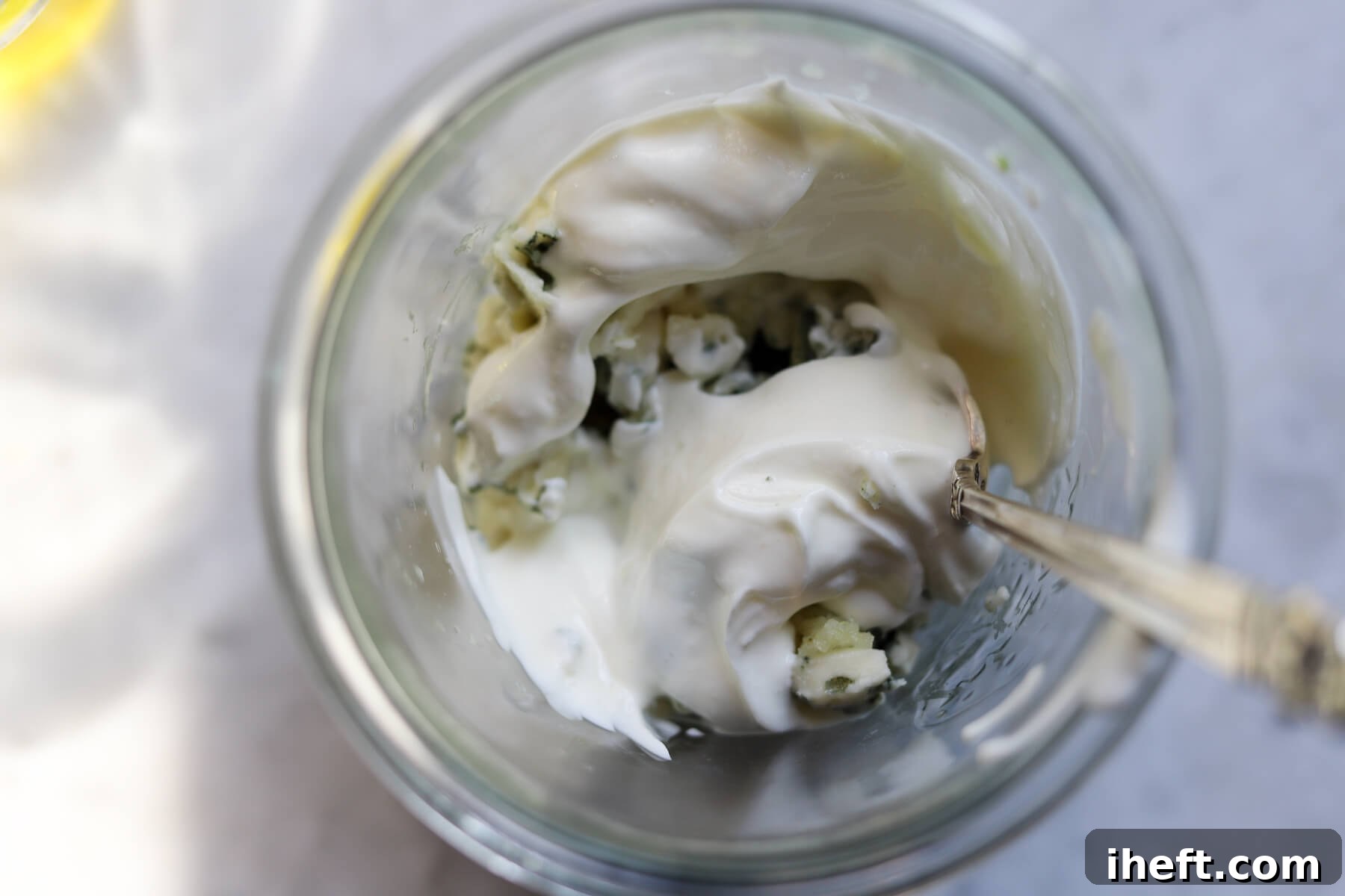 Blue cheese, yogurt, and garlic are stirred together in a glass jar with a silver spoon.