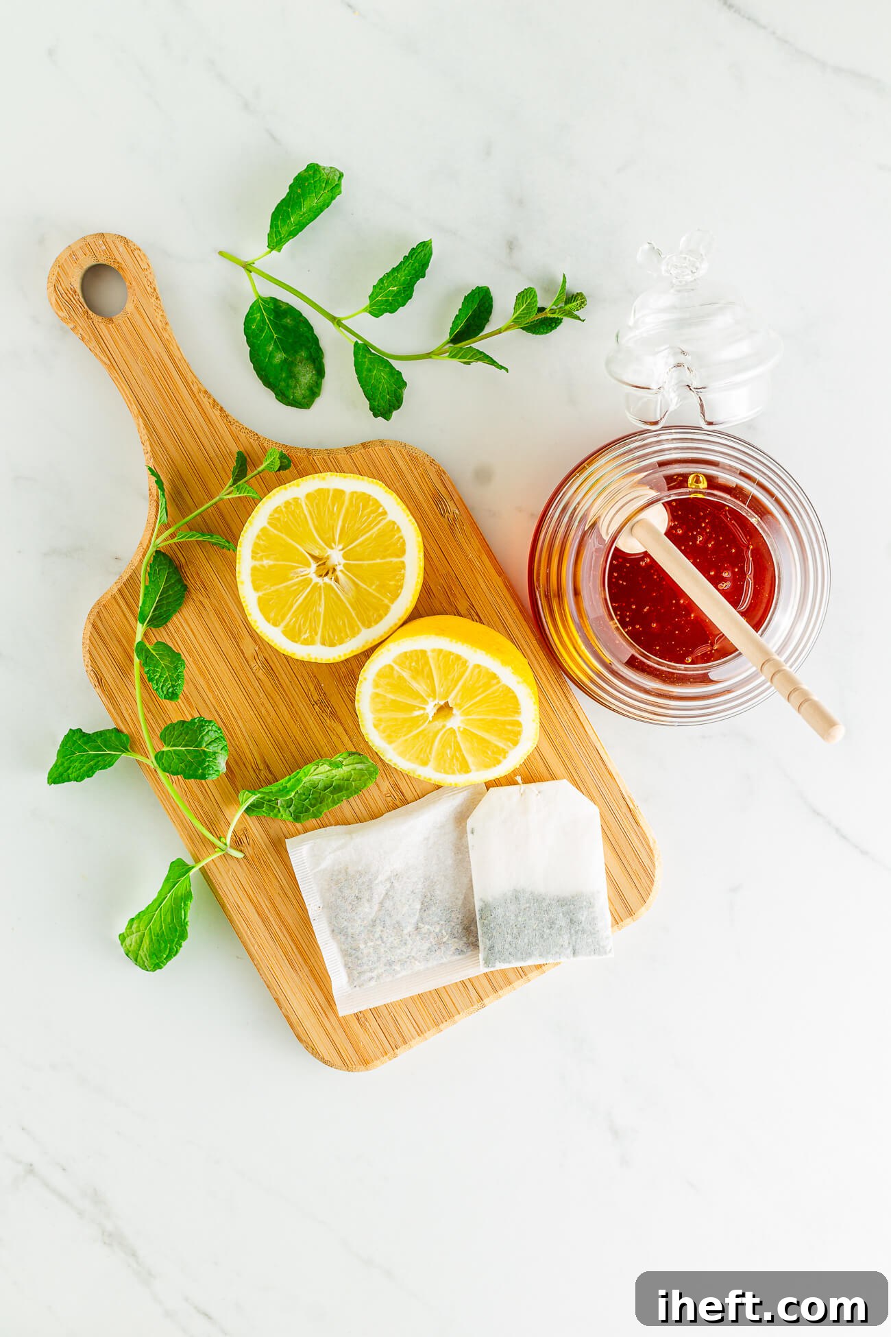 A lemon cut in half, fresh mint sprigs, two tea bags, and honey on a marble counter. Essential ingredients for a homemade Medicine Ball.