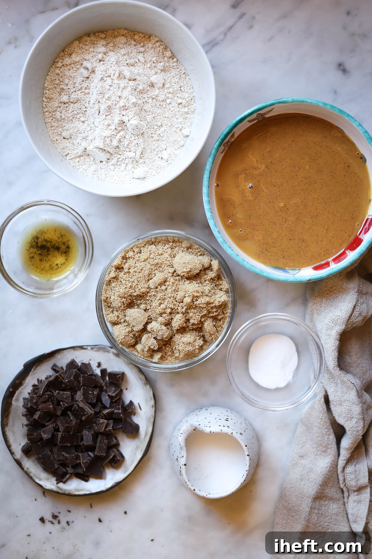 The essential ingredients for Billie Eilish's oat flour peanut butter cookies are neatly arranged on a pristine marble countertop. Bowls contain oat flour, creamy peanut butter, rich brown sugar, baking soda, almond milk, pure vanilla extract, and finely chopped dark chocolate.