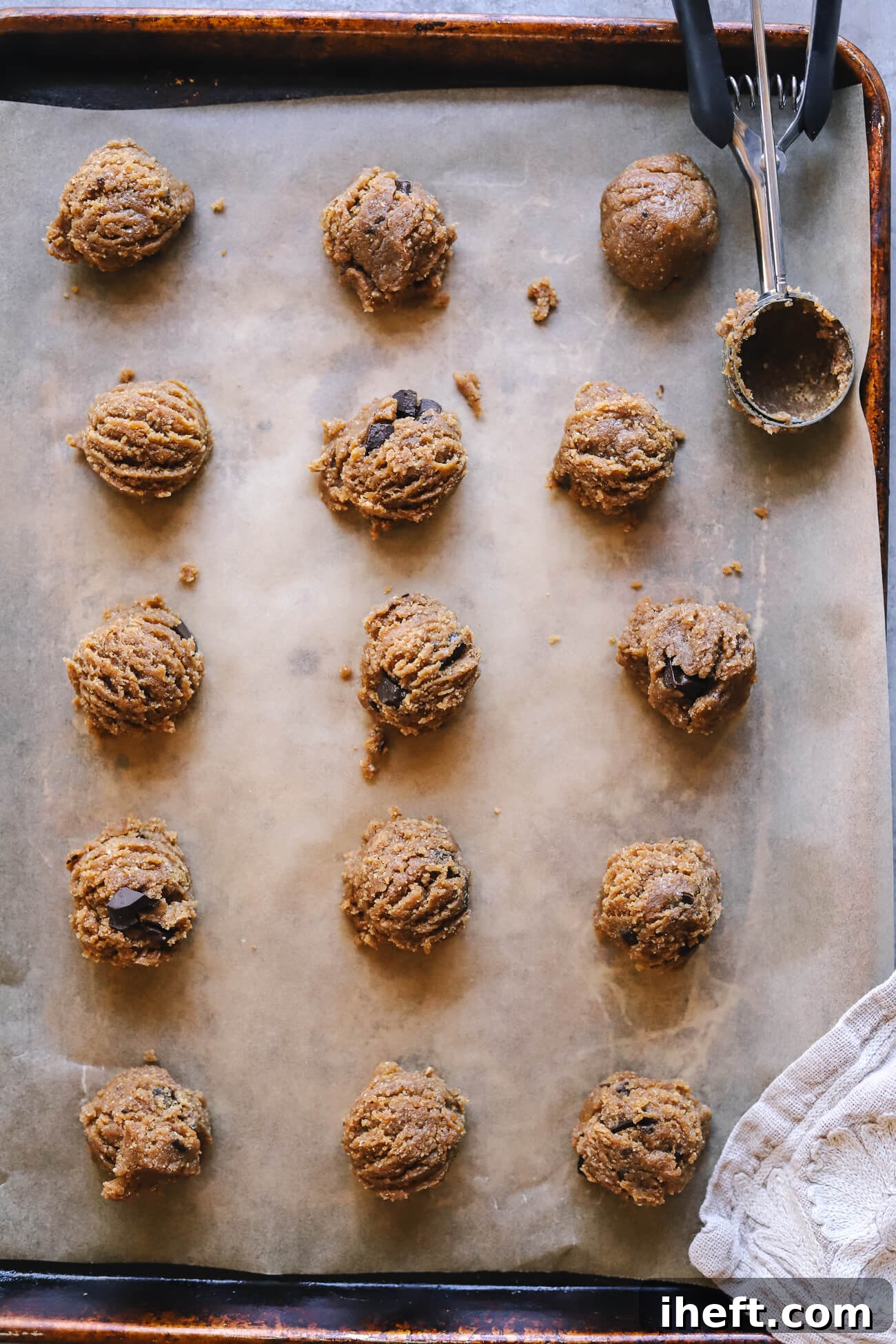 Fifteen perfectly scooped balls of vegan gluten-free peanut butter cookie dough are neatly arranged on a parchment-lined cookie sheet, awaiting their bake. A cookie scoop rests in the upper right corner, ready for more.