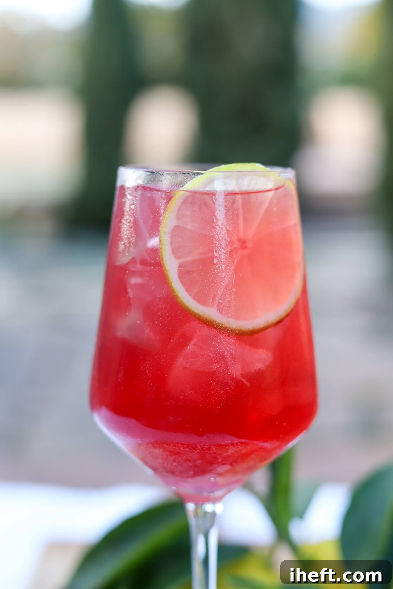 A close-up photo of a vibrant red sparkling Sleepy Girl Mocktail beverage with ice and a fresh lime wheel, elegantly presented in a wine glass.