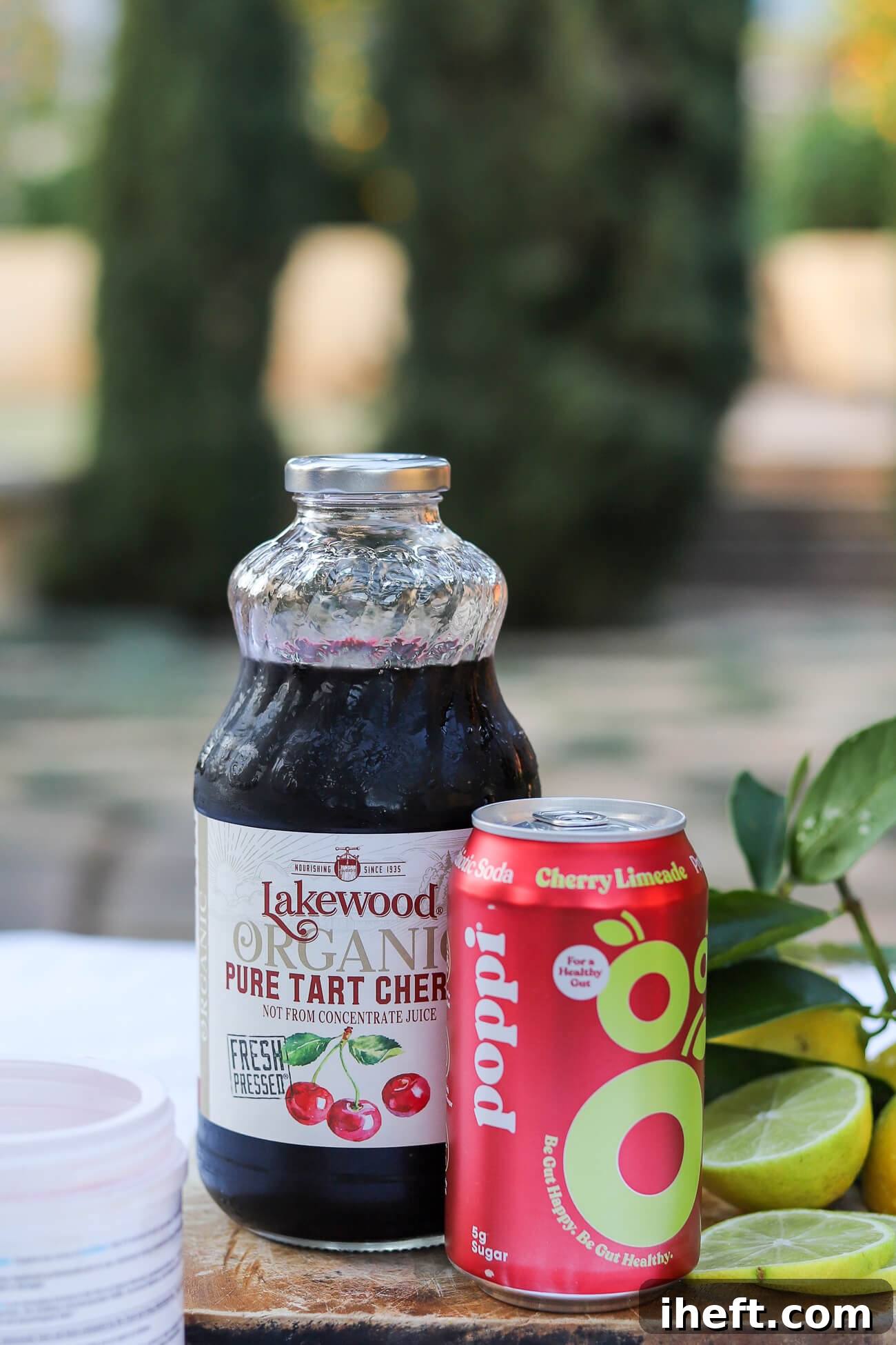 A curated display of Sleepy Girl Mocktail ingredients on a wooden board: a bottle of pure tart cherry juice, a can of prebiotic soda, a tub of magnesium powder, and fresh limes, all set outdoors.