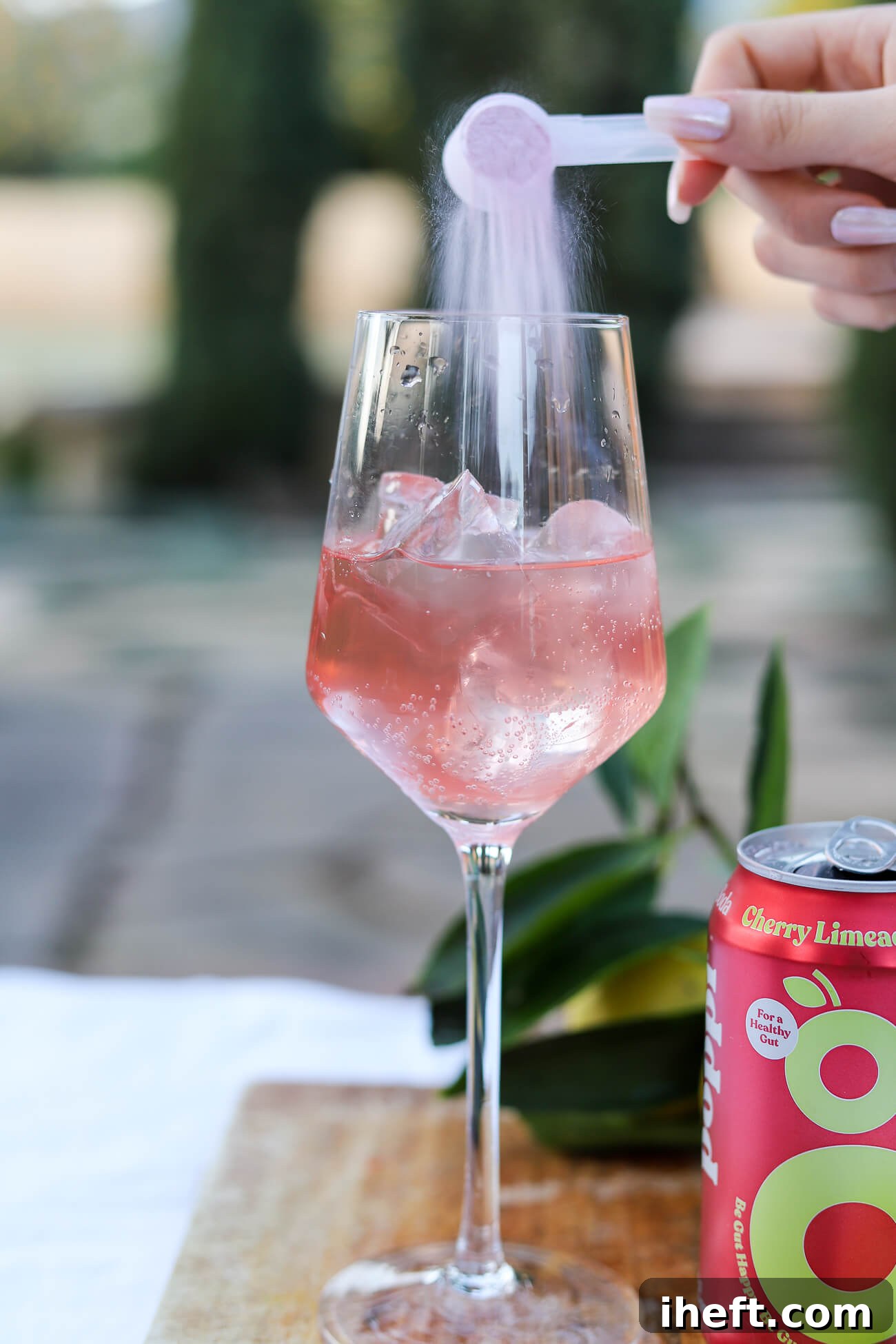 A hand gently pours a scoop of light pink magnesium powder into a wine glass filled with ice and sparkling prebiotic soda, creating a soothing magnesium drink.