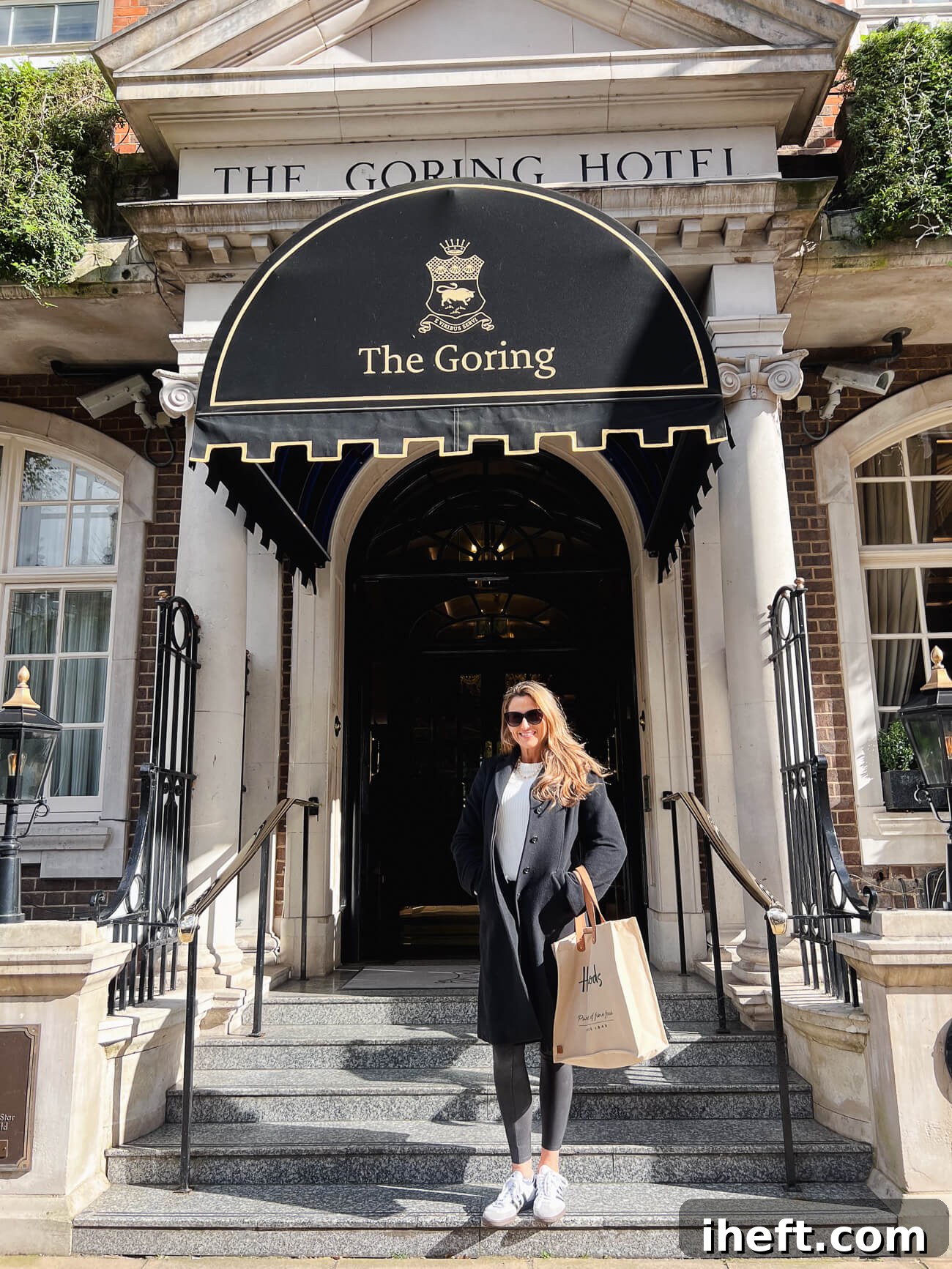 Marina stands on the steps in front of The Goring, a luxury hotel in Belgravia, London. She holds a Harrods shopping bag and wears a black overcoat. 
