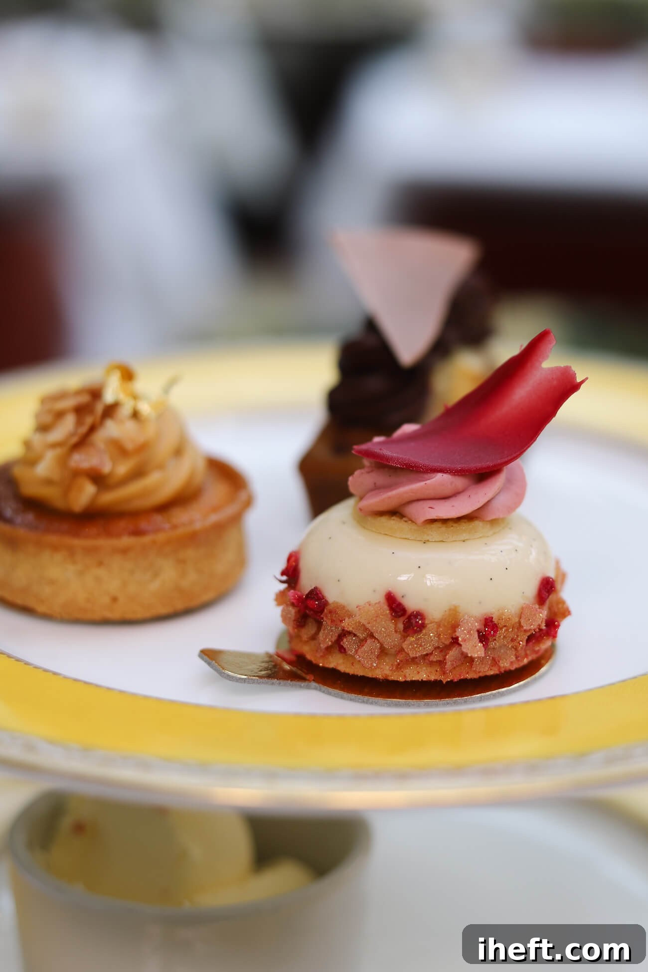 Three small fancy afternoon tea desserts sit on the top tier of a tea stand at the Goring. In the foreground is a vanilla bean panna cotta with pink garnishes. In the background is an apple tart and a chocolate tart. 