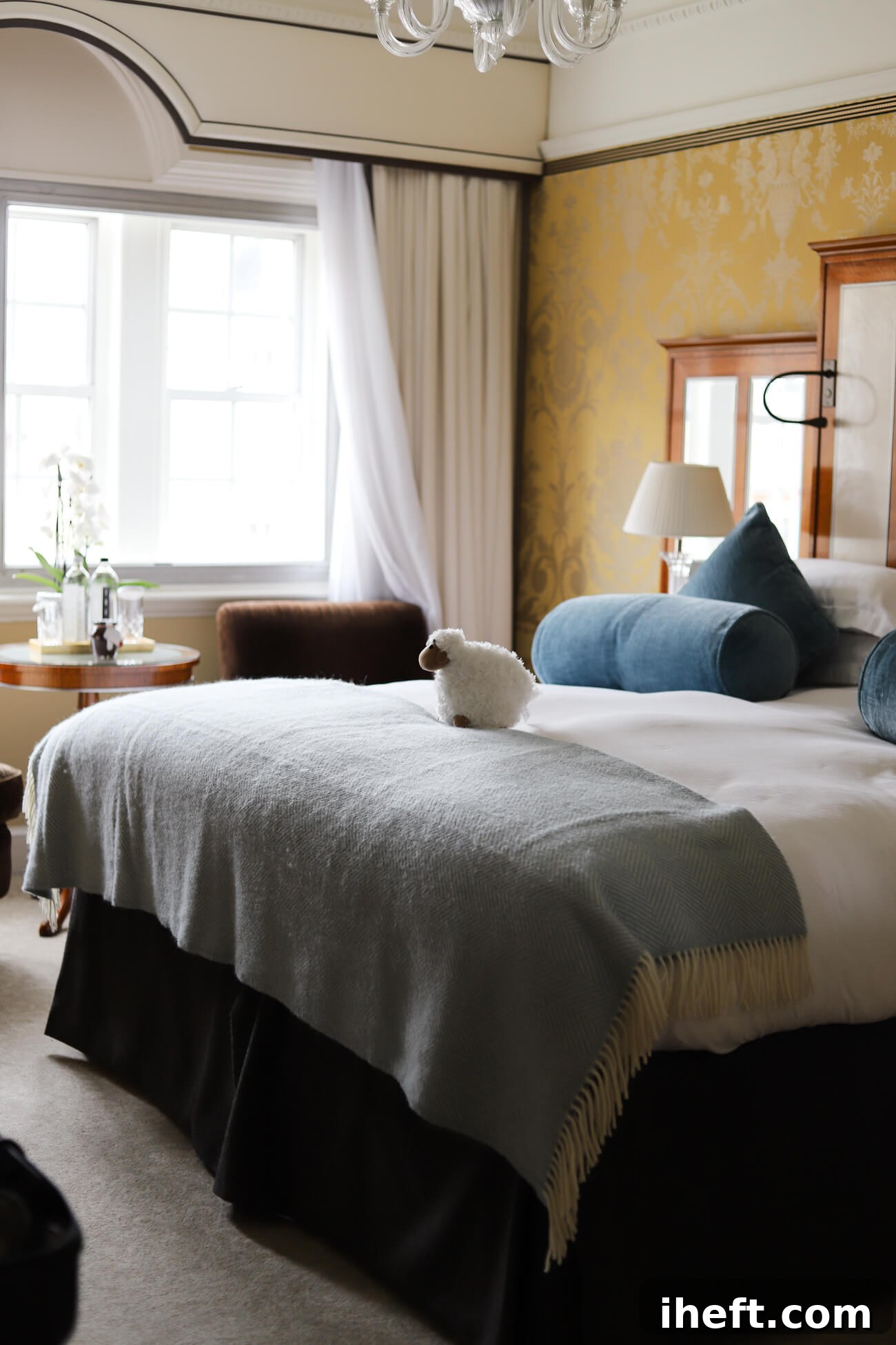 A superior room at the Goring hotel. A queen bed with white linens topped with a blue blanket and throw pillows. A small stuffed sheep sits on top of the bed. A chair and side table can be seen in the background. 