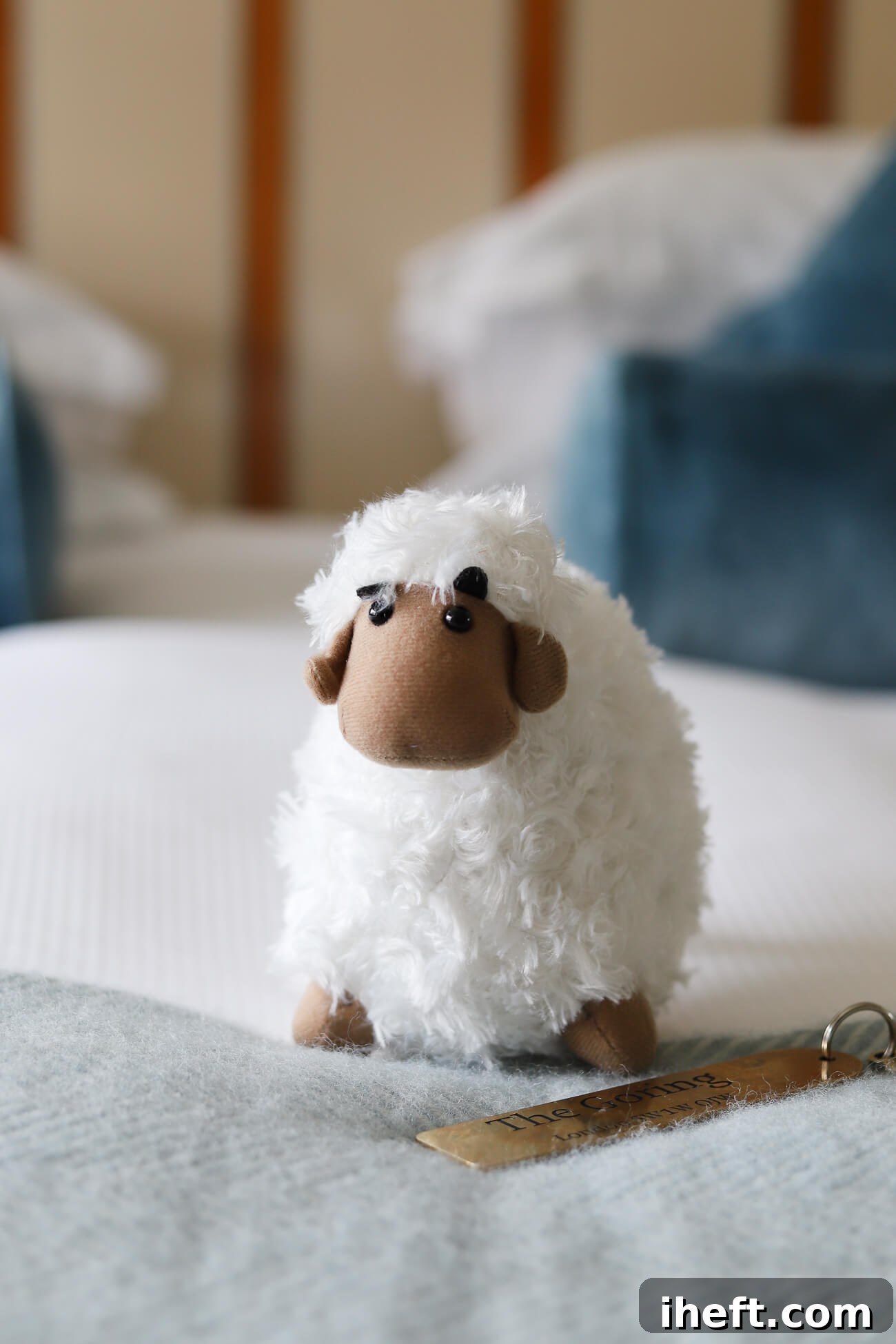 A cute plush sheep sits on a bed made with white and blue linens. A key with "The Goring" engraved is next to the sheep. 