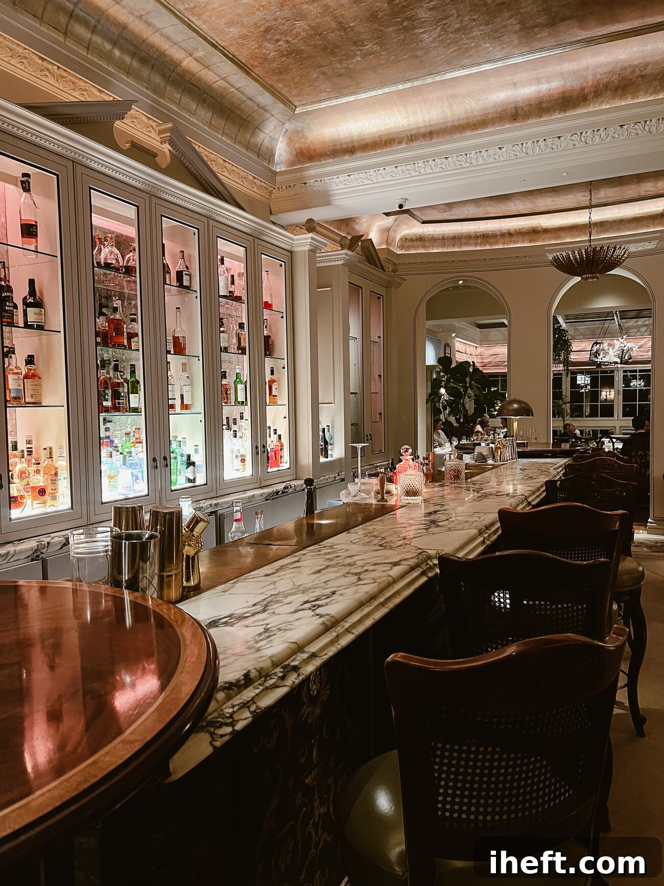 A long marble counter with dark wood barstools in front of white cabinets holding a variety of alcohols in the Goring hotel bar. 
