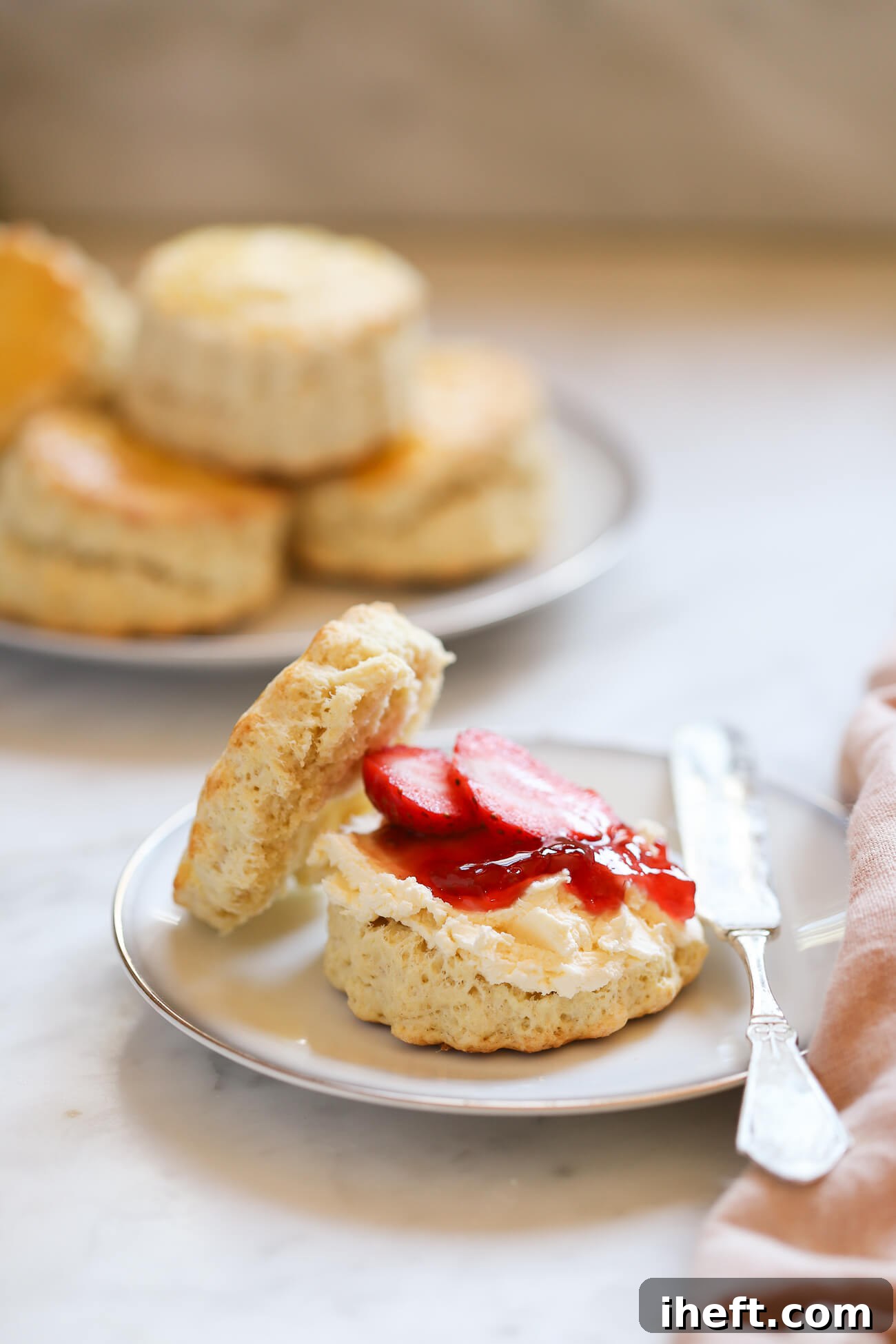 An authentic English scone topped with a generous dollop of clotted cream and a spoonful of strawberry jam, garnished with fresh strawberry slices. A plate of more freshly baked classic scones is softly blurred in the background.