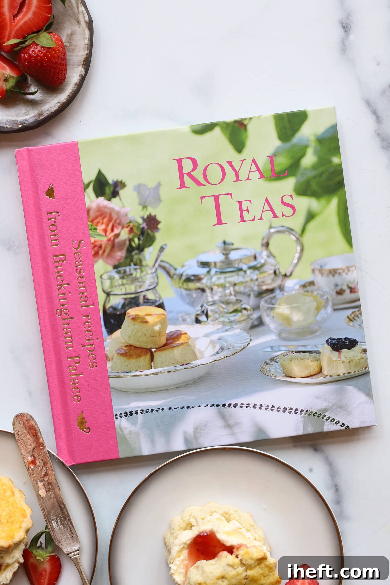 A charming cookbook with a pink spine, featuring a cover photo of beautifully arranged scones with clotted cream, jam, and a teacup on an outdoor table. The title reads "Royal Teas: Seasonal recipes from Buckingham Palace." 