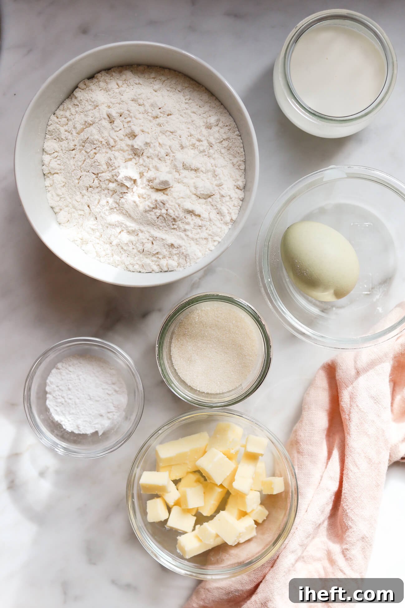 A collection of fresh ingredients for English scones artfully arranged in bowls on a pristine marble countertop: all-purpose flour, fresh milk, a single egg, granulated sugar, precisely diced cold butter, and baking powder.