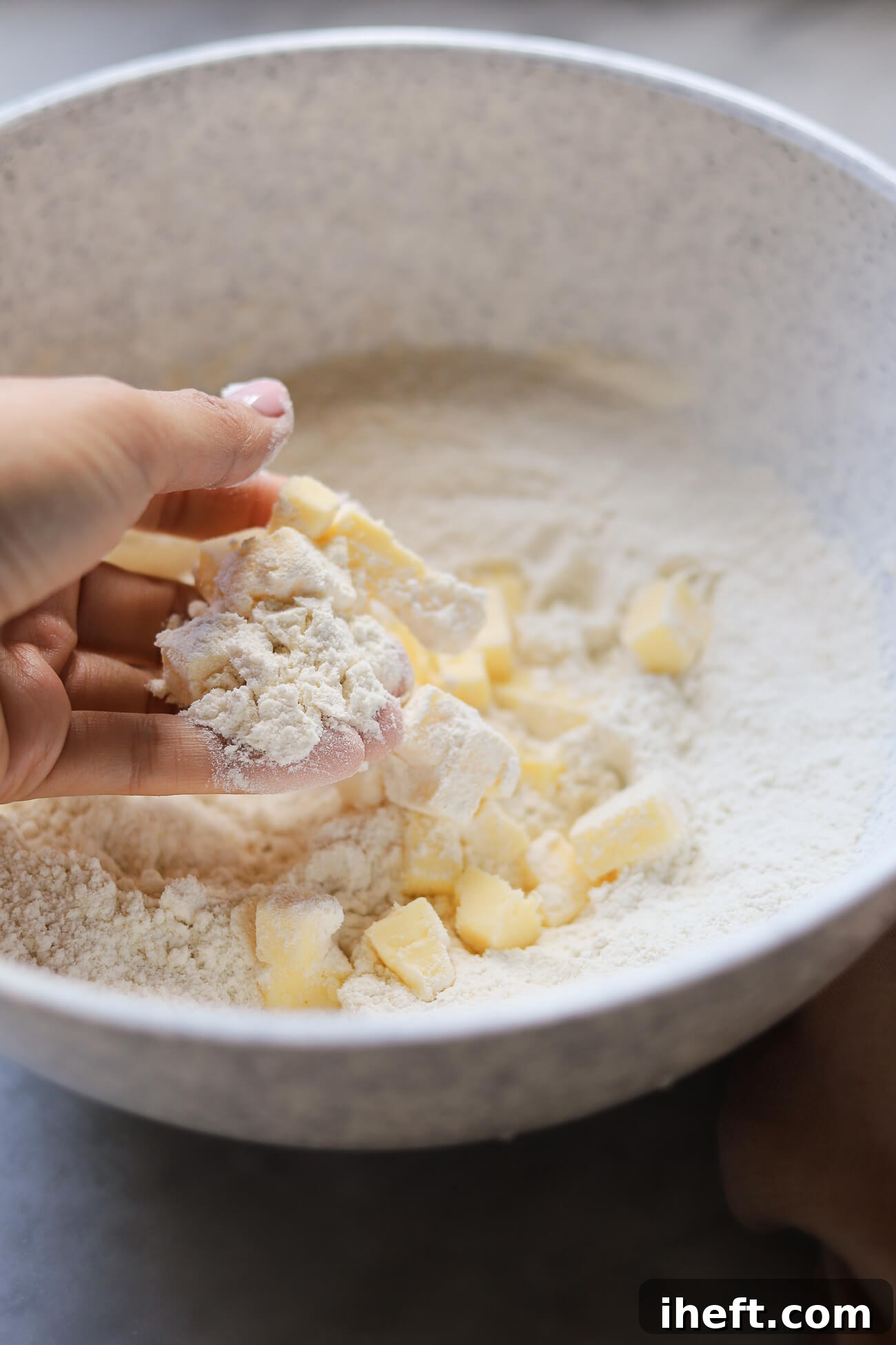 Diced cold butter is being carefully rubbed into a mixture of dry ingredients (flour, baking powder, sugar, salt) using fingertips on a marble countertop, preparing the base for English scones.
