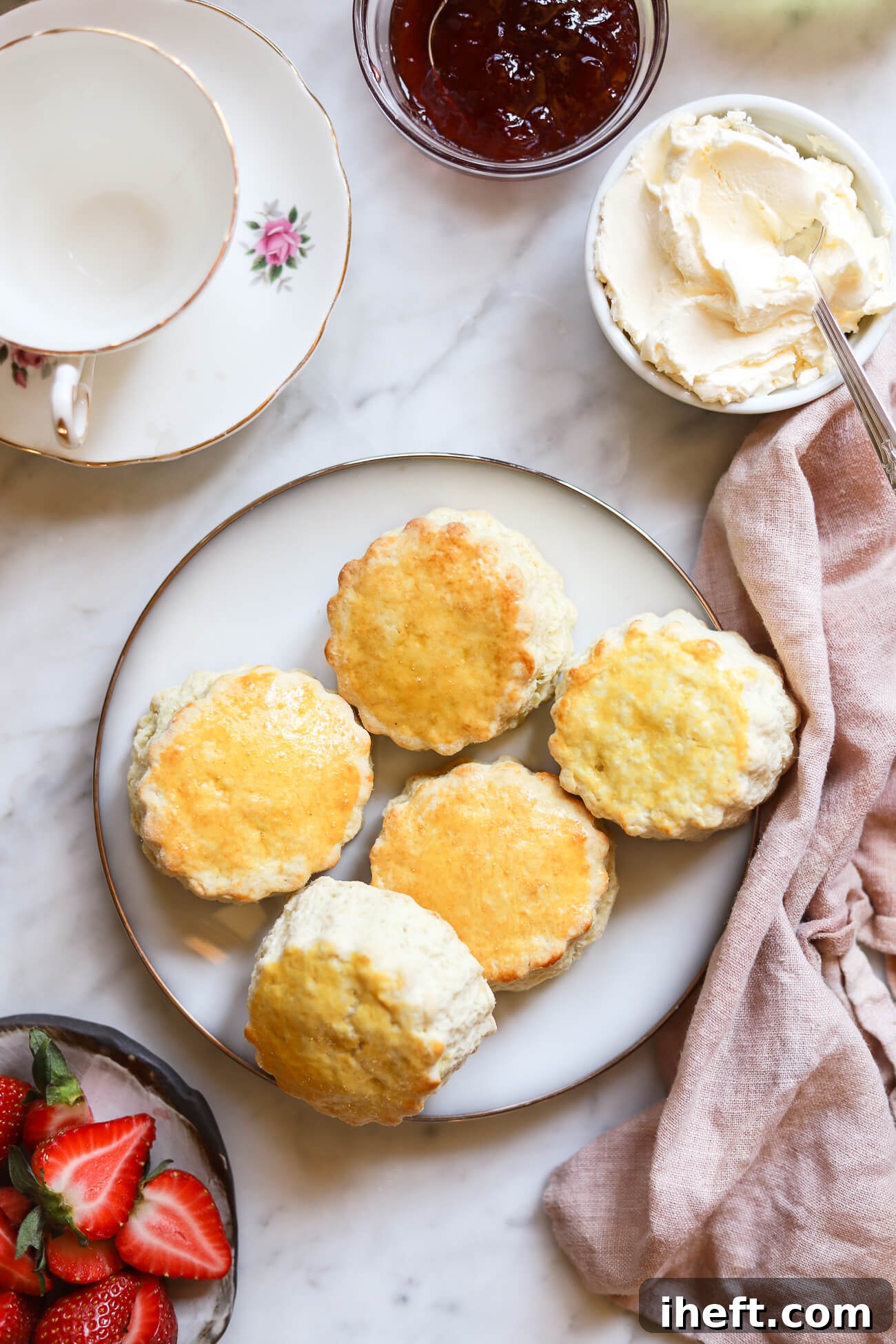 A pristine white plate holds five freshly baked, perfectly golden British scones. Surrounding them are small, elegant bowls brimming with rich clotted cream and vibrant strawberry jam, alongside a scattering of fresh strawberries and a delicate English teacup, all set on a smooth countertop.