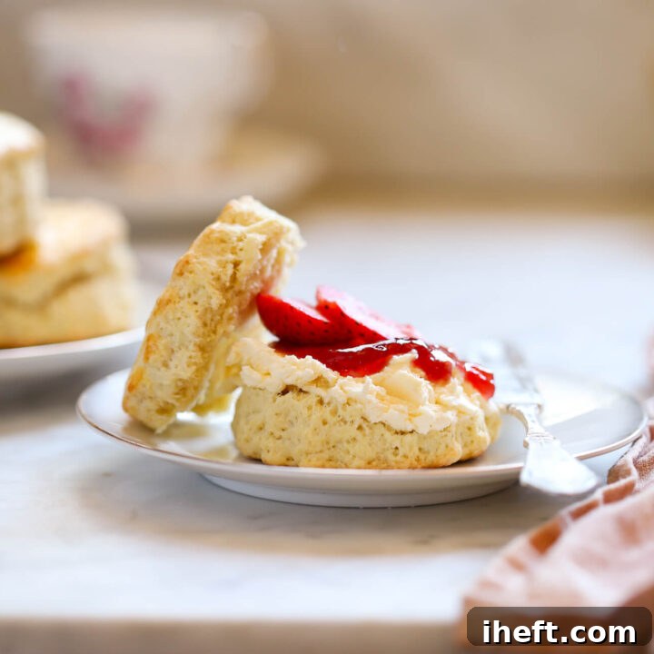 A traditional English scone, beautifully split and generously adorned with clotted cream and vibrant strawberry jam, rests on a small white plate. In the background, three more plain, freshly baked British scones are arranged on another plate.