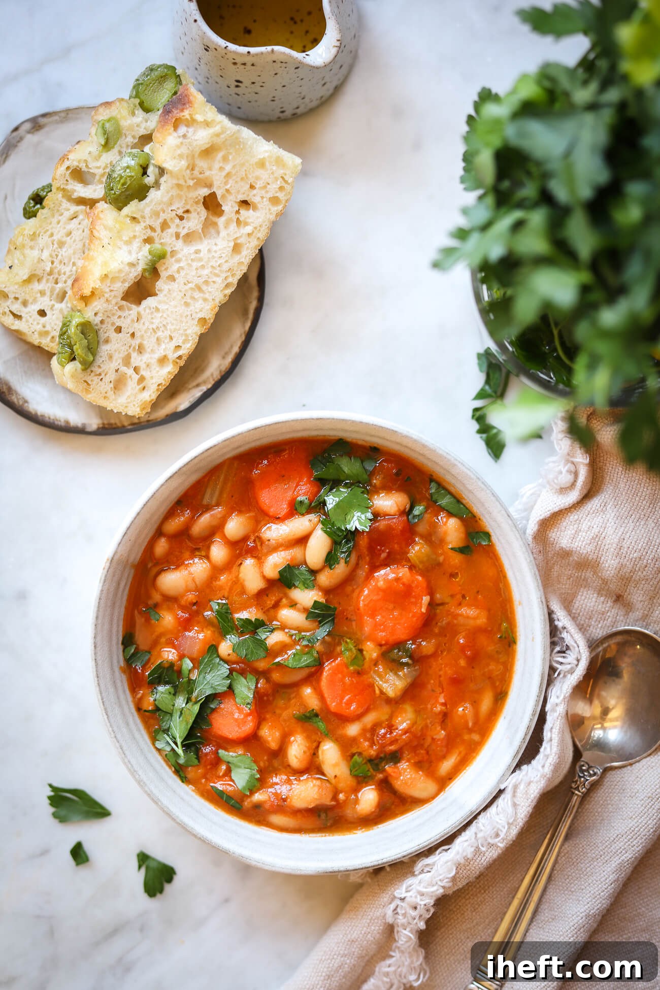 An overhead food photo of Greek white bean soup with carrots and parsley in a light grey ceramic bowl. Olive focaccia and parsley are on the side. 