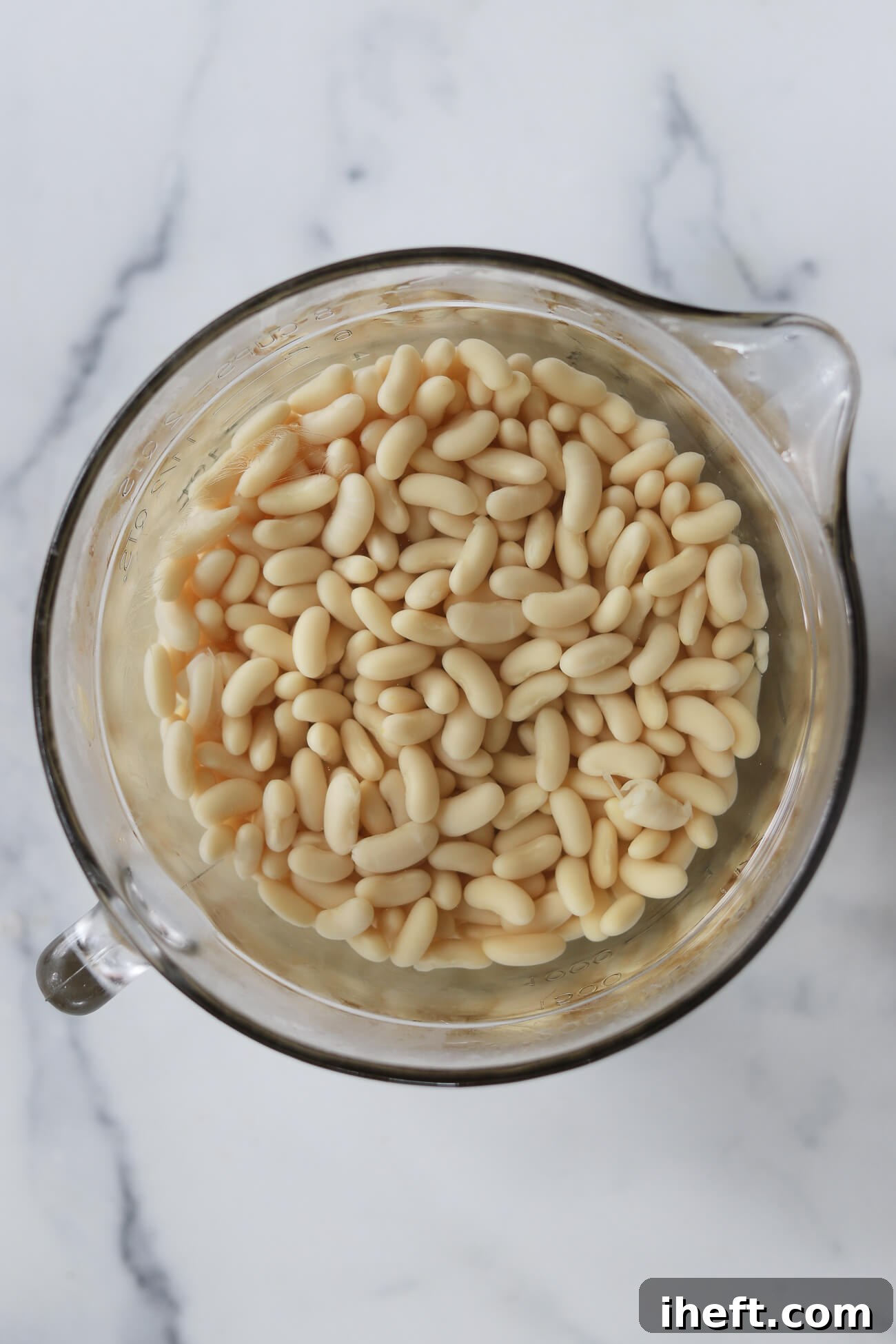 Dried cannellini beans soaking in a glass bowl of water on a marble countertop before making Greek bean soup. 