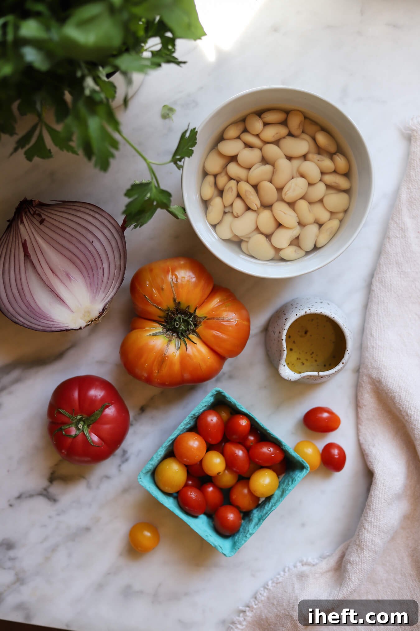 A bowl of cooked butter beans in a white bowl alongside a small dish of olive oil, cherry and heriloom tomatoes, and a red onion on a white marble countertop. Fresh parsley and basil are in the corner. These are ingredients for a healthy vegetarian butter bean salad recipe. 