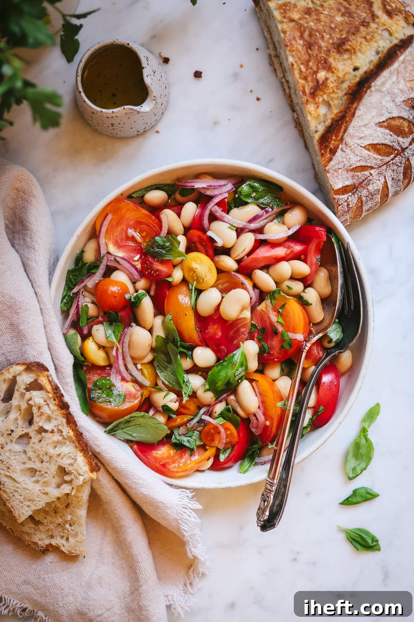 Beautiful overhead food photography of a white rimmed plate filled with heirloom tomato, butter bean, and basil salad with silver serving utensils. The salad sits on a marble counter with a small dish of dressing and loaf of homemade sourdough on the side. 