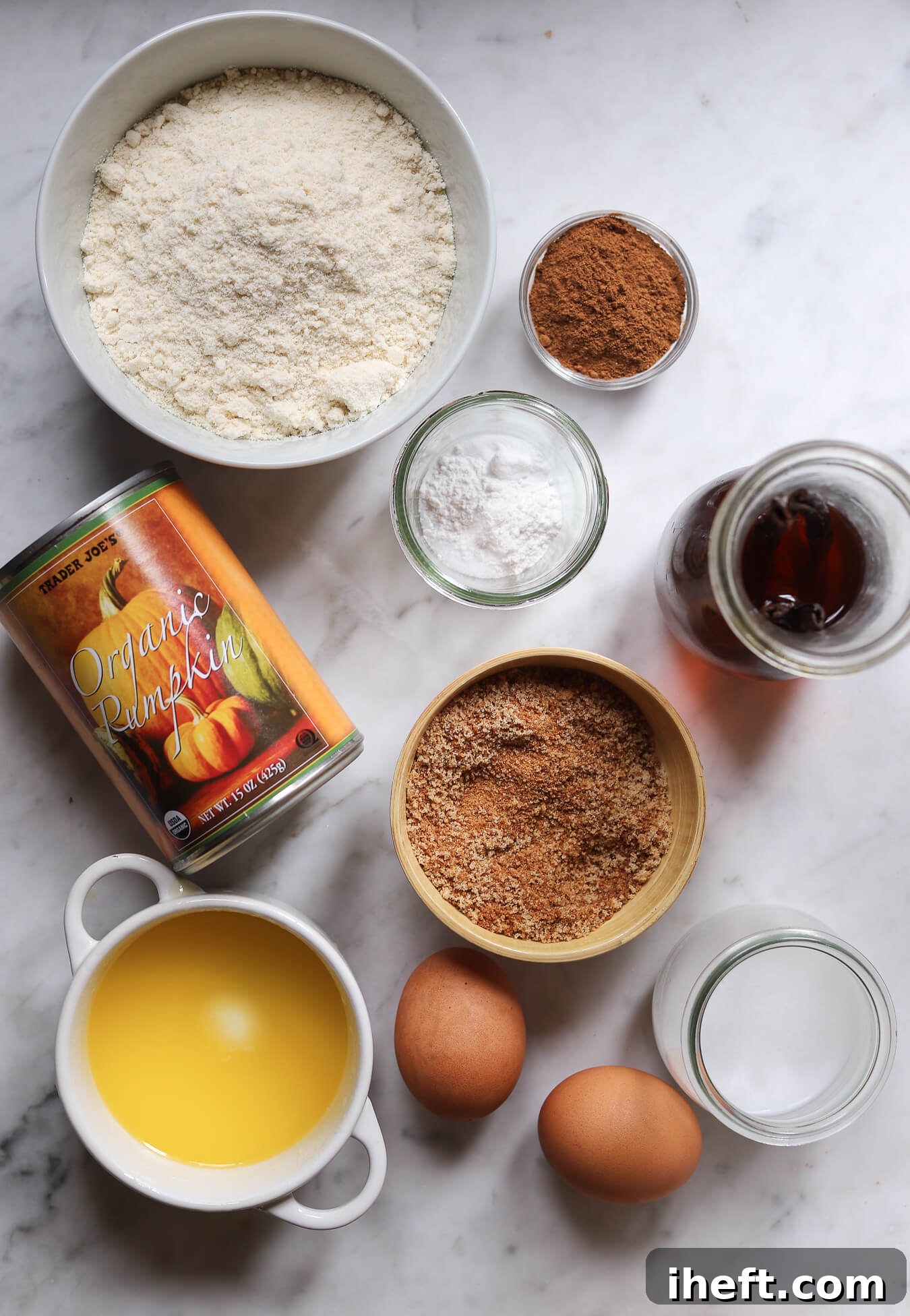 The ingredients for gluten free pumpkin bread on a white marble counter: almond flour, pumpkin pie spice, baking soda and baking powder, vanilla extract, coconut sugar, eggs, milk, melted butter and canned organic pumpkin. 