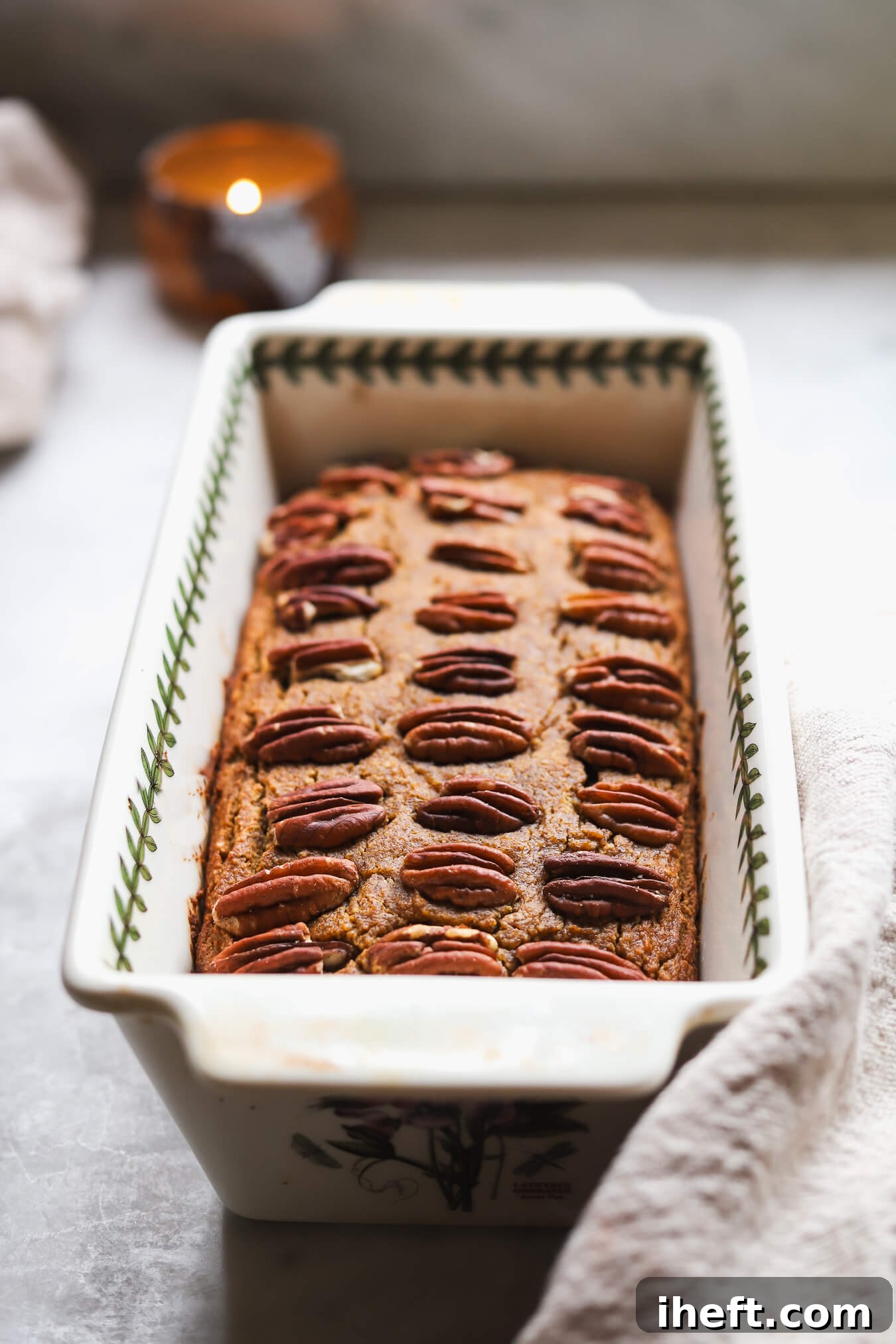 A loaf of gluten free, grain free, paleo pumpkin bread made with almond flour topped with pecan halves in a porcelain loaf pan on a marble kitchen counter. This healthy pumpkin bread looks delicious! 