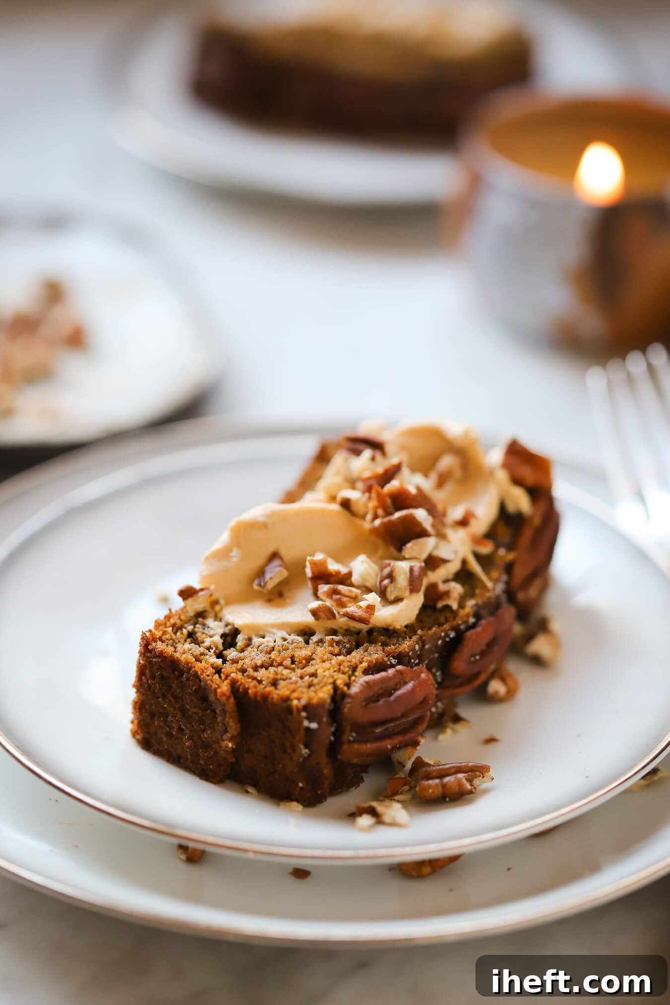 A slice of gluten free pumpkin bread on a white plate with a candle in the background. The bread slice is topped with pumpkin cream cheese and pecans. 
