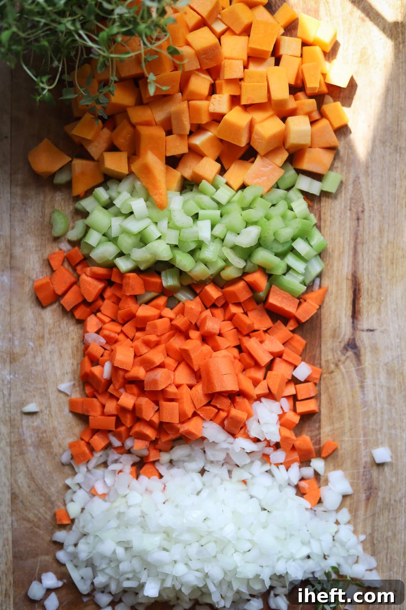 Chopped butternut squash, celery, carrots, and onion with fresh thyme on a cutting board. 