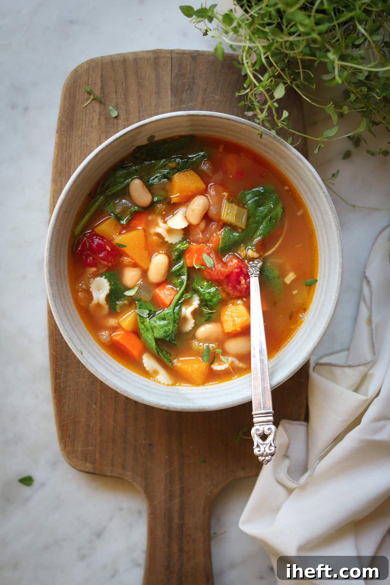 An overhead photo of a ceramic bowl filled with Ina Garten's Winter Minestrone made with butternut squash, white beans, pasta, and spinach. Fresh thyme is in the corner. 