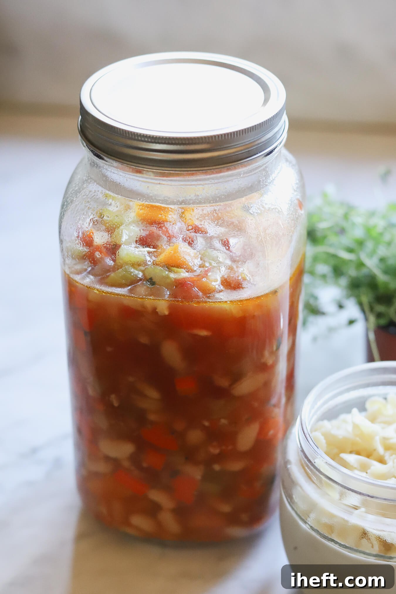 A large mason jar filled with minestrone soup and a separate bowl of cooked small pasta to make Winter Minestrone ahead of time. 