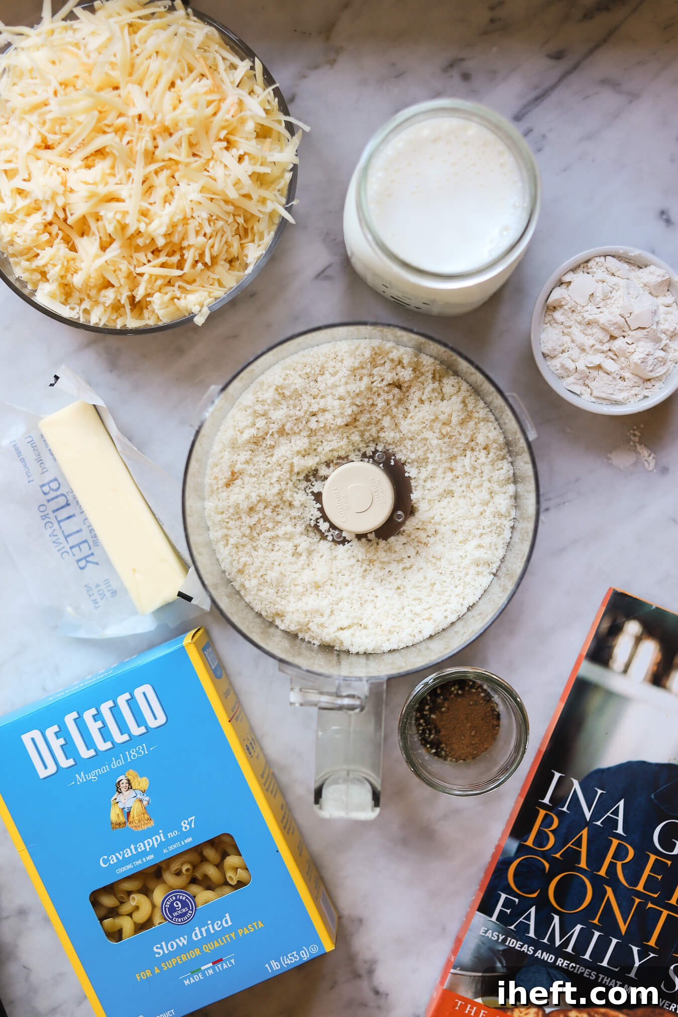 The ingredients for baked mac and cheese on a white marble countertop. These include, shredded cheeses, milk, flour, breadcrumbs, butter, and pasta. 