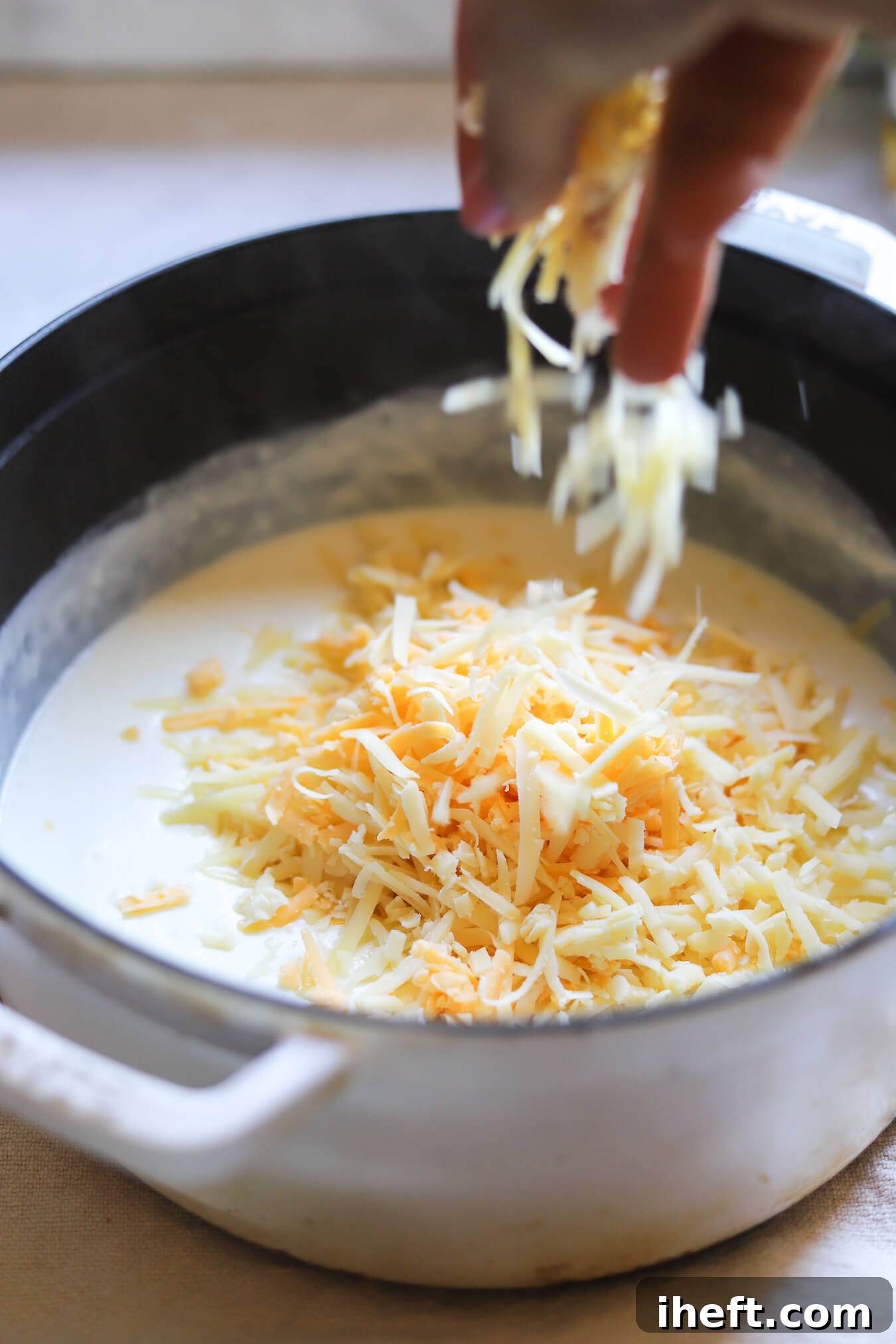 Shredded gruyere and cheddar cheeses are added to a pot to make sauce for baked mac and cheese. 
