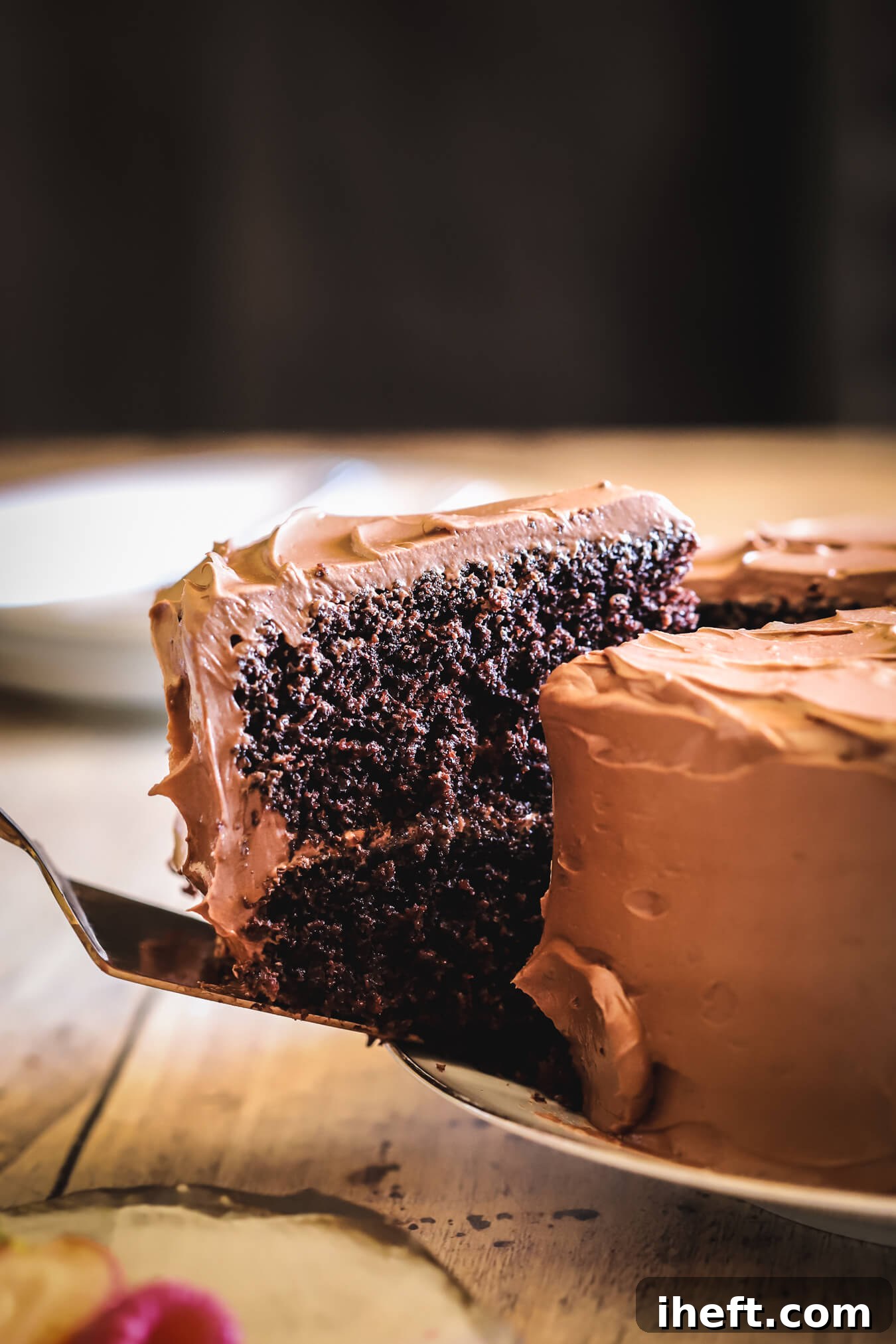 A elegant gold cake utensil delicately lifts a perfect slice of Beatty's Chocolate Cake from a white plate, showcasing its moist layers and rich frosting.