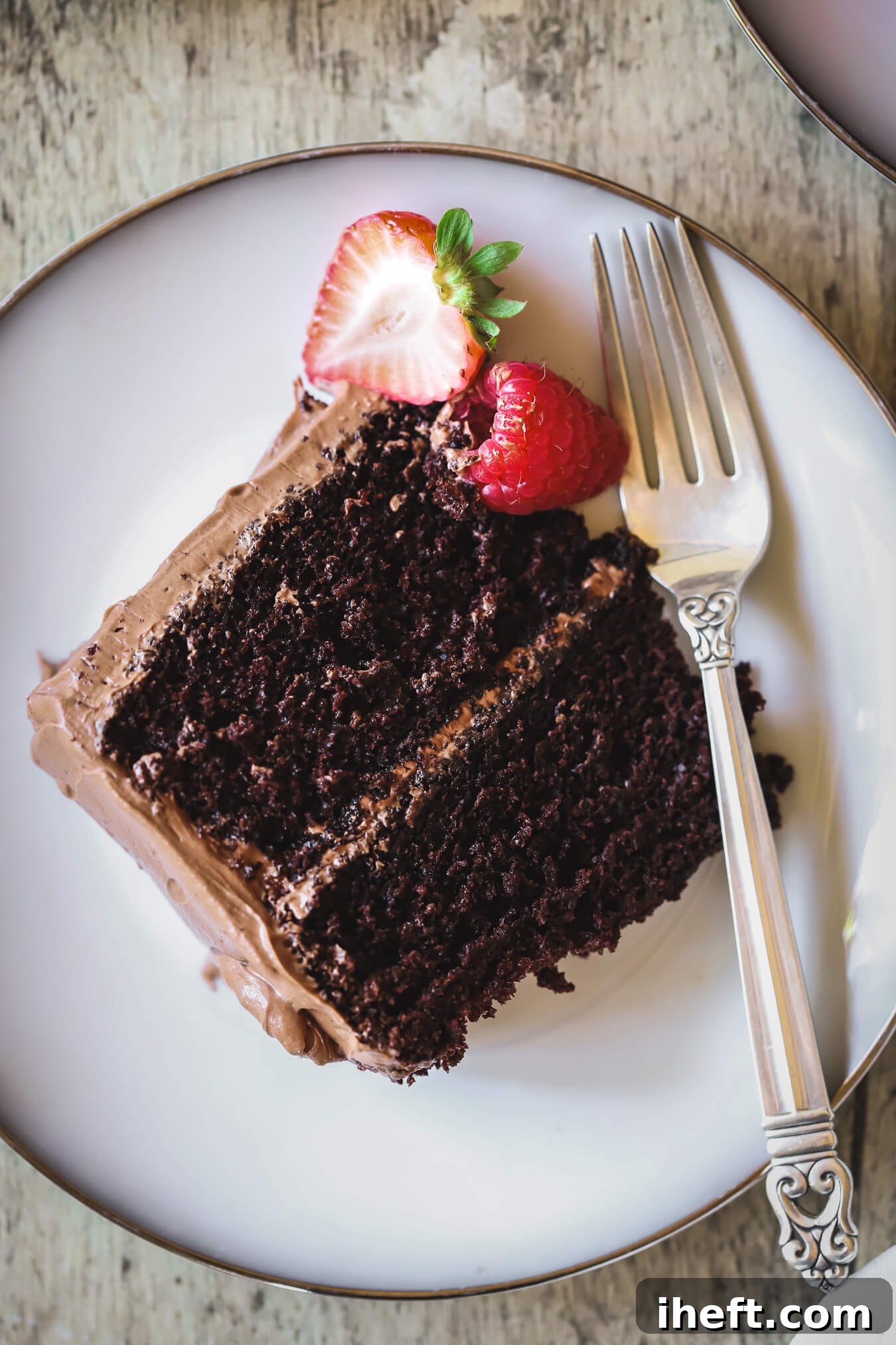 An inviting overhead shot of a generous slice of Ina Garten's Beatty's Chocolate Cake, presented on a pristine white plate with a silver fork, a fresh strawberry half, and a vibrant raspberry. This delicious, moist two-layer chocolate cake is crowned with luscious chocolate buttercream.