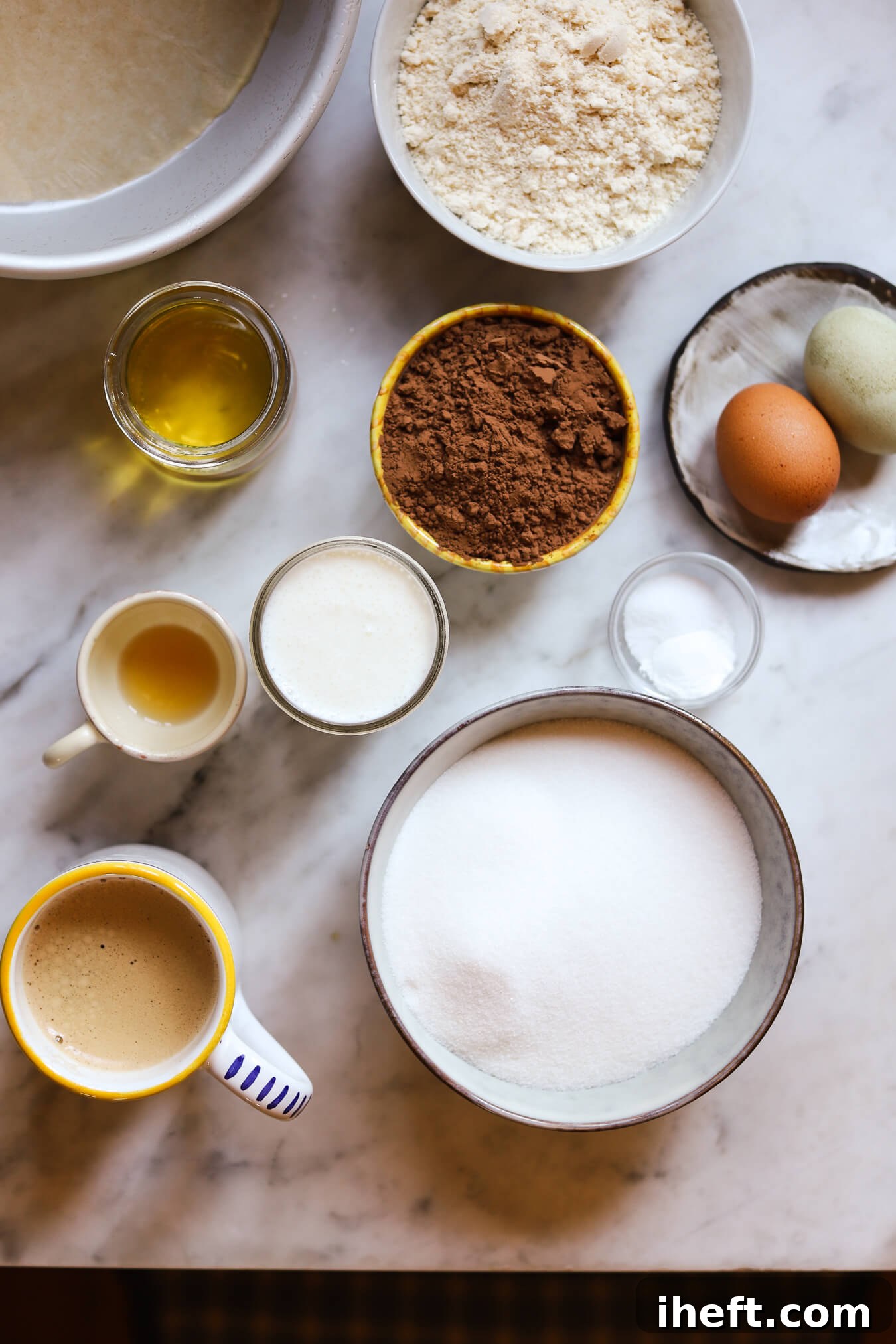 A curated collection of ingredients for Ina Garten's chocolate cake, artfully arranged in bowls on a pristine white marble kitchen counter. Featuring flour, two fresh eggs, baking powder and soda, rich cocoa powder, creamy buttermilk, granulated sugar, pure vanilla extract, a neutral oil, and fragrant coffee.