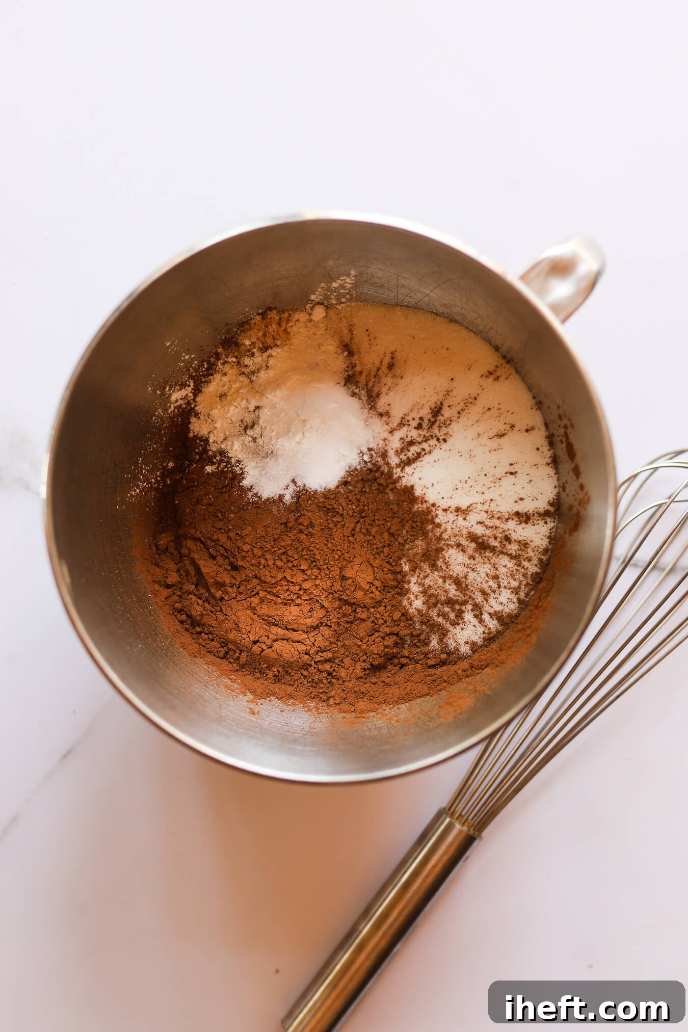 The initial dry ingredients for Beatty's Chocolate Cake, including sugar, flour, cocoa, baking soda, baking powder, and salt, are expertly whisked together in the gleaming bowl of a KitchenAid stand mixer, ready for the next step.